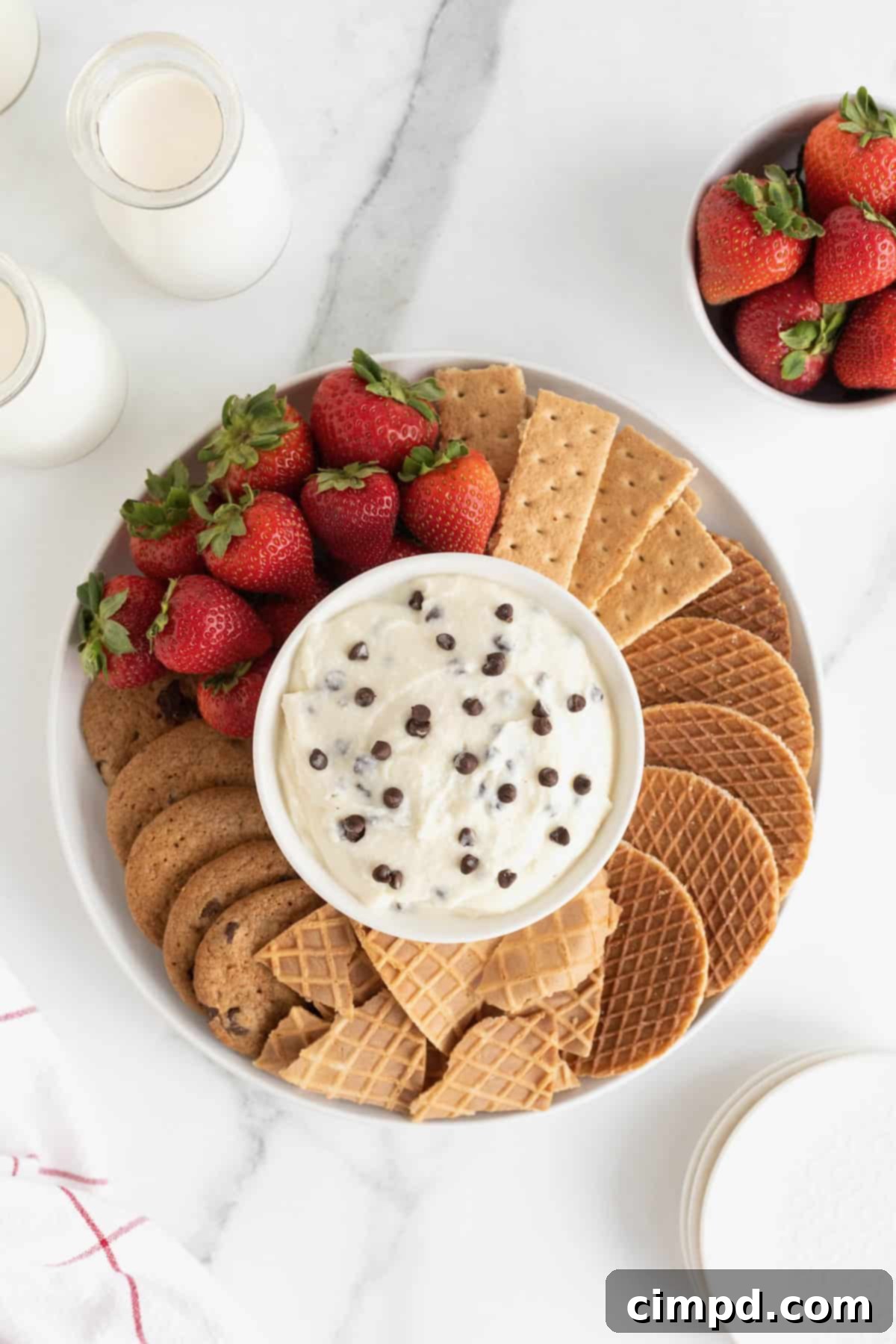 A white serving platter filled with cookies, graham crackers, strawberries and sugar cone wafers. In the center of the dish is a white bowl filled with chocolate chip cannoli dip.