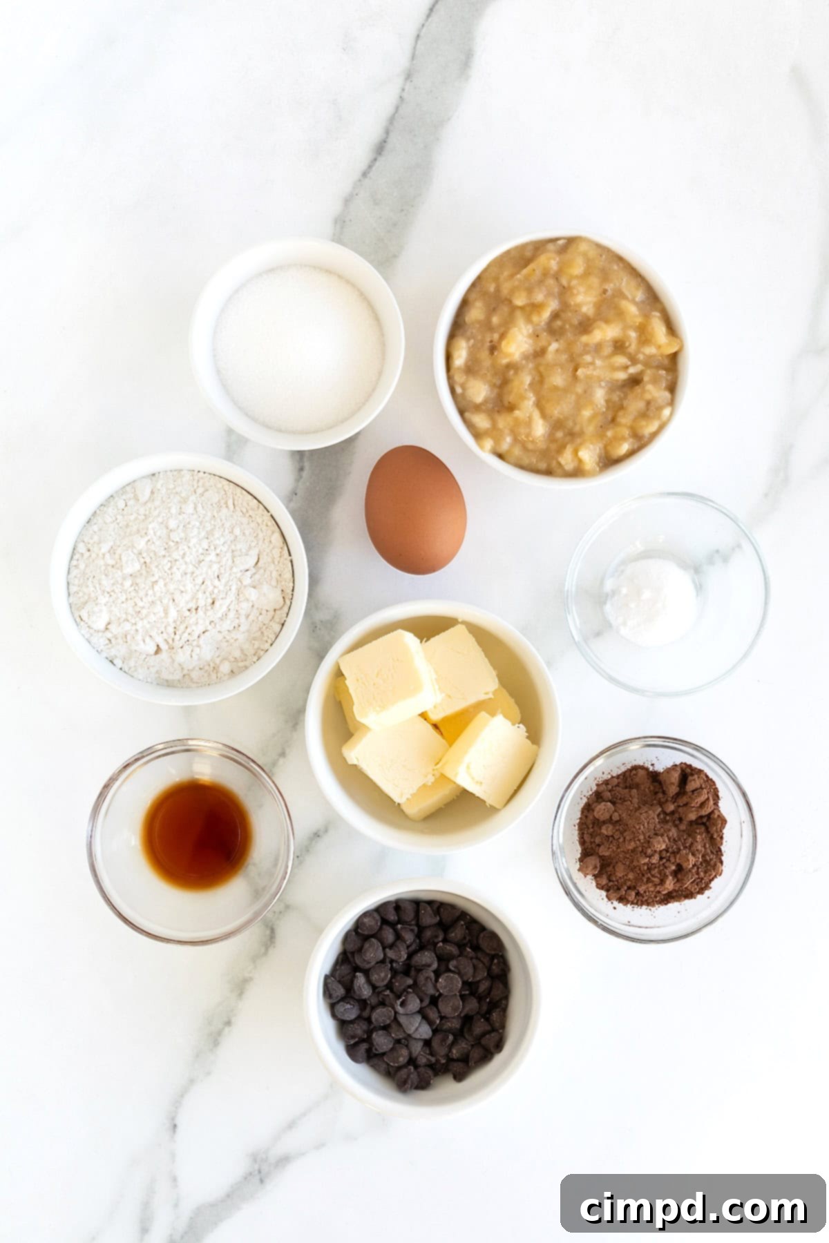 Chocolate Banana Dream Muffins 5 All the fresh ingredients for making double chocolate banana muffins neatly arranged in small white glass dishes on a pristine white marble counter, ready for preparation.