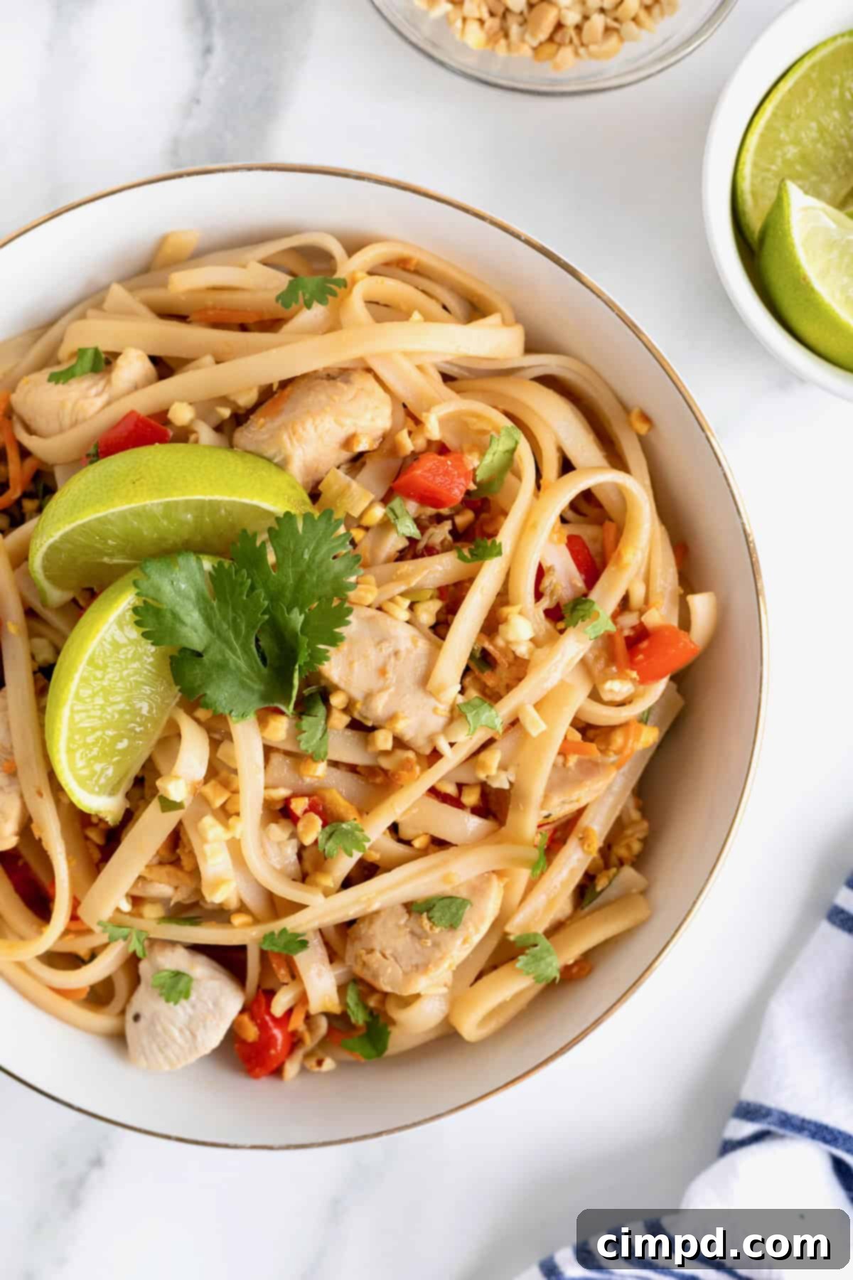 A white porcelain bowl filled with chicken pad Thai, elegantly garnished with a bright green lime wedge and fresh cilantro leaves, presented on a pristine white marble counter.