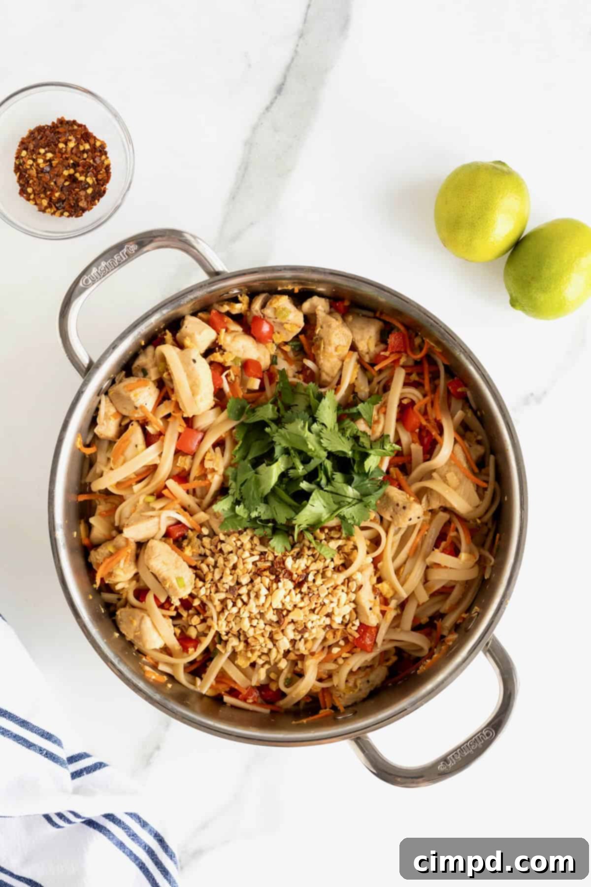 A stainless steel pot brimming with chicken pad Thai, artfully garnished with vibrant fresh cilantro and generously sprinkled chopped peanuts, all set on a sophisticated white marble counter.