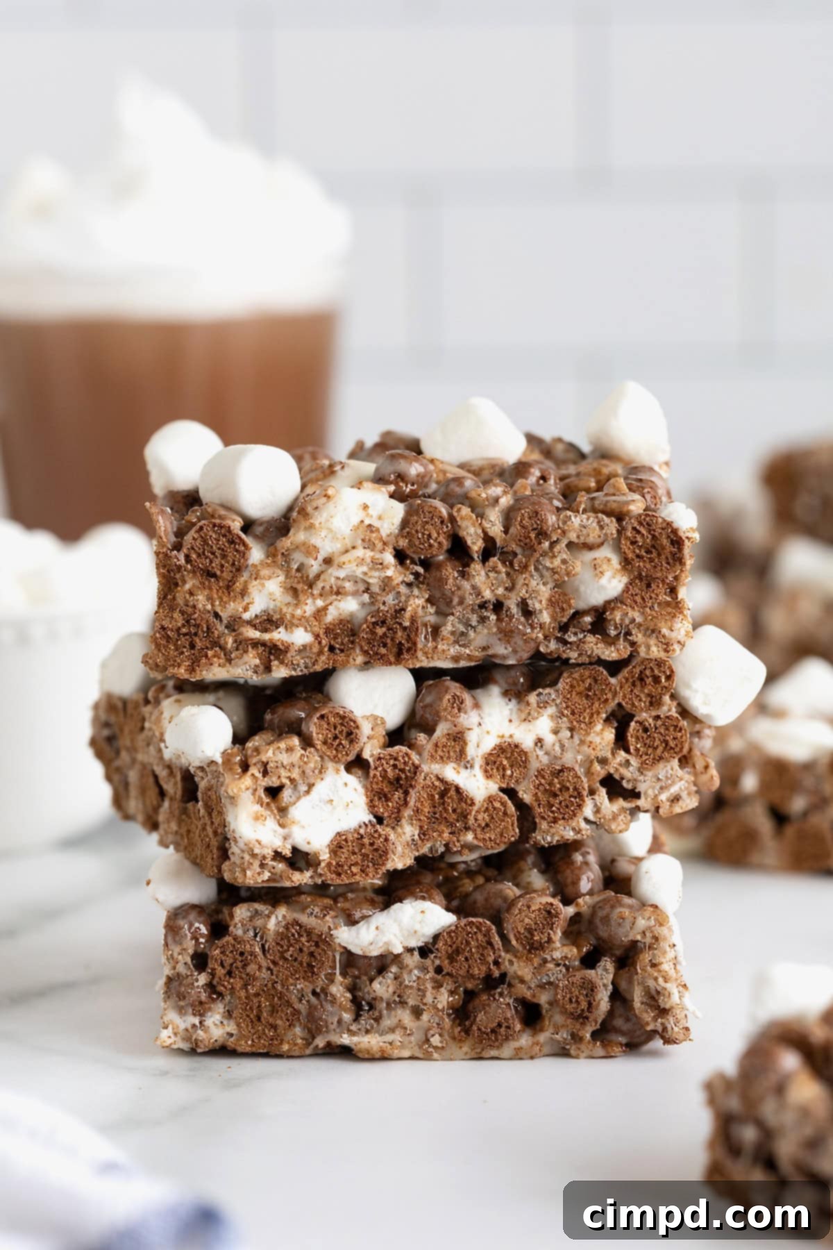 Cozy Hot Cocoa Krispies Squares 2 Three no-bake hot cocoa Krispie treats topped with mini marshmallows stacked on a white surface. A glass mug of hot cocoa sits in the background.