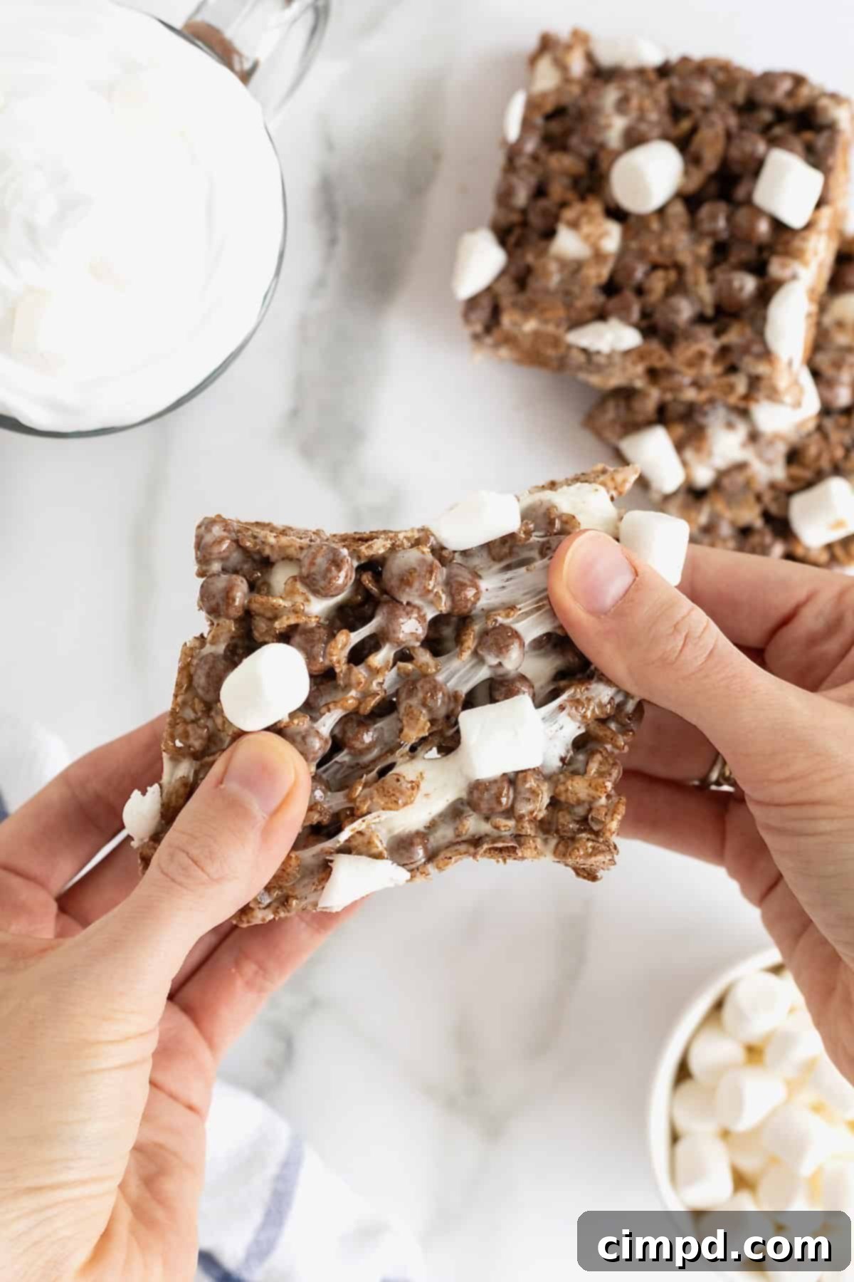 Cozy Hot Cocoa Krispies Squares 3 A pair of hands pulling apart a chocolate marshmallow cereal treat, showcasing its delightfully chewy, gooey texture.