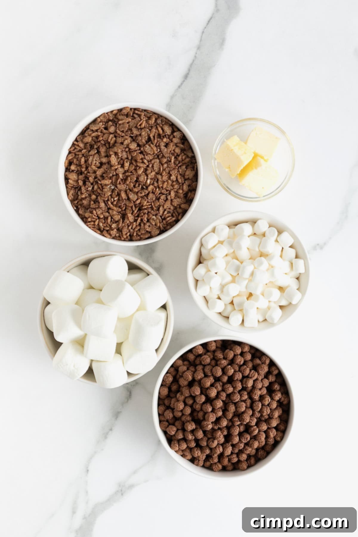 Cozy Hot Cocoa Krispies Squares 5 Ingredients for Hot Cocoa Krispies Treats laid out in small white dishes on a white marble counter, showing butter, marshmallows, Cocoa Puffs, and Cocoa Krispies.