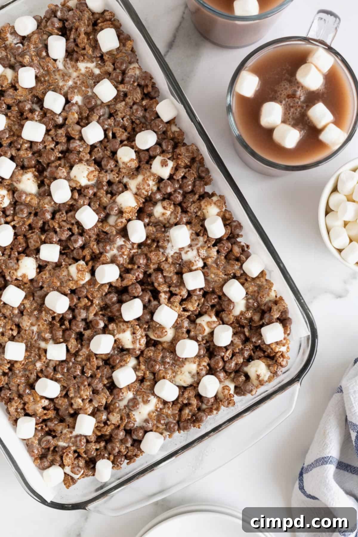 Cozy Hot Cocoa Krispies Squares 9 A glass baking dish of hot cocoa Krispie treats, perfectly cut and ready to serve, on a white marble counter, showing their appealing texture and mini marshmallow topping.