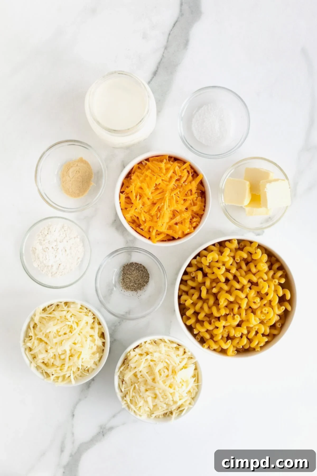Ingredients for baked Mac and Cheese arranged in small glass dishes on a pristine white marble counter.