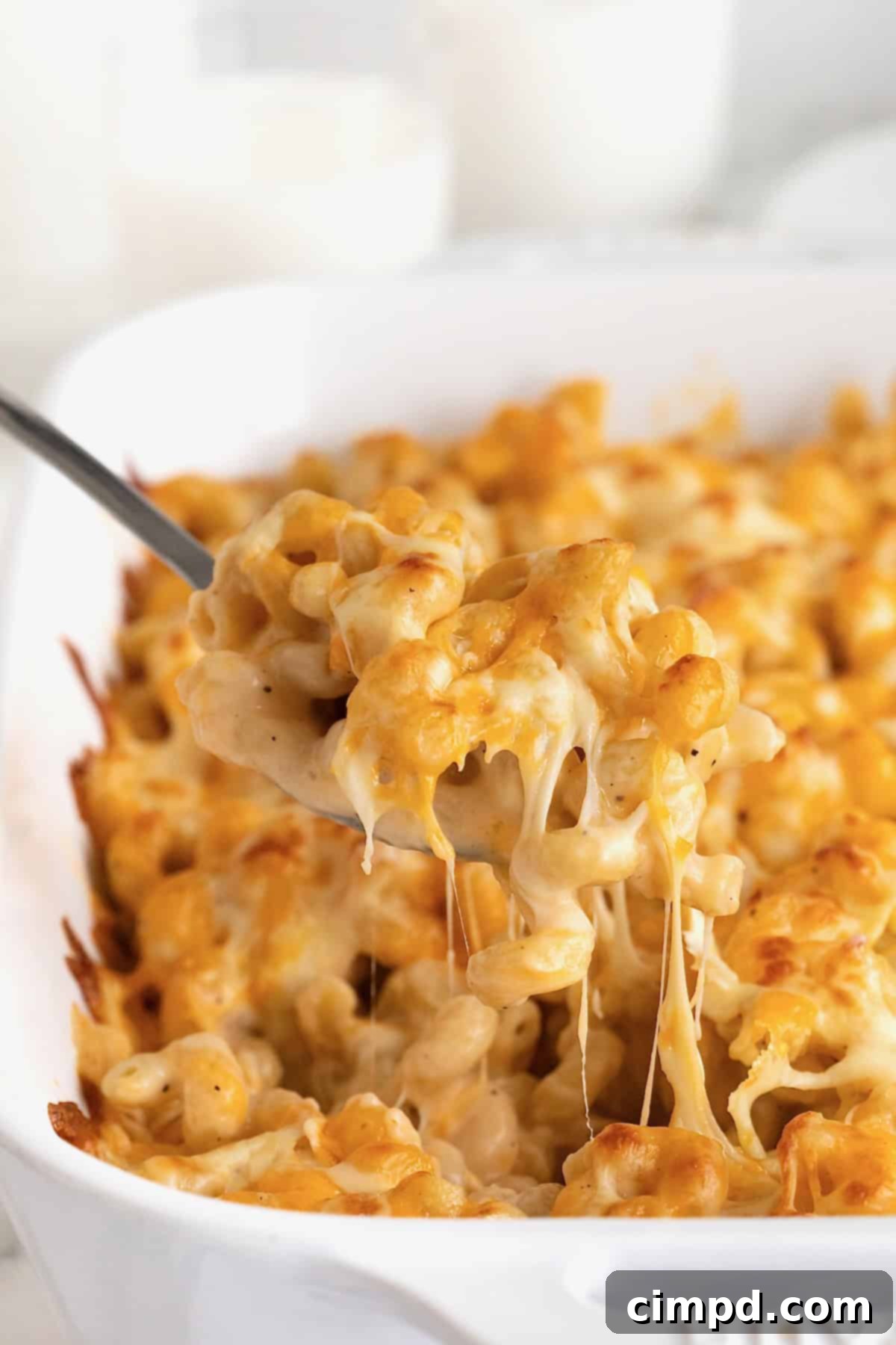 A spoon lifting a spoonful of Mac and cheese out of a white ceramic baking dish, showing the gooey cheese.
