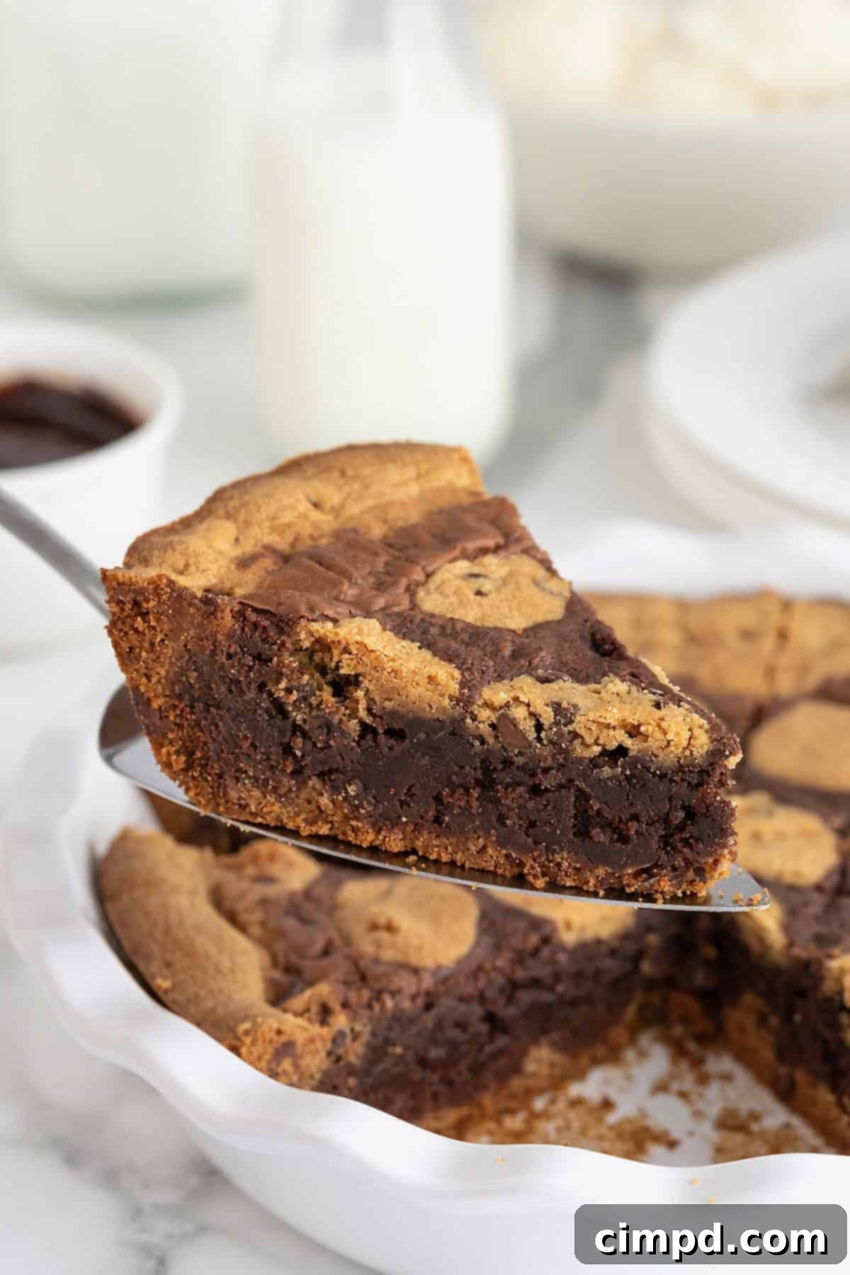 A slice of brookie pie being lifted from a white porcelain pie plate on a metal spatula. The pie features a golden cookie crust, a dark chocolate brownie layer, and chunks of baked cookie dough on top.