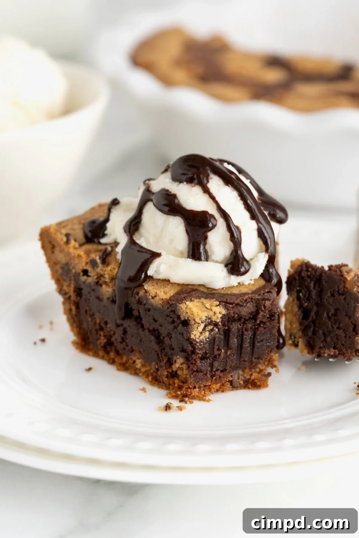 A slice of brookie pie topped with a scoop of melting vanilla ice cream and rich chocolate syrup on a small white plate. A bite has been taken from the pie, revealing its layered texture.