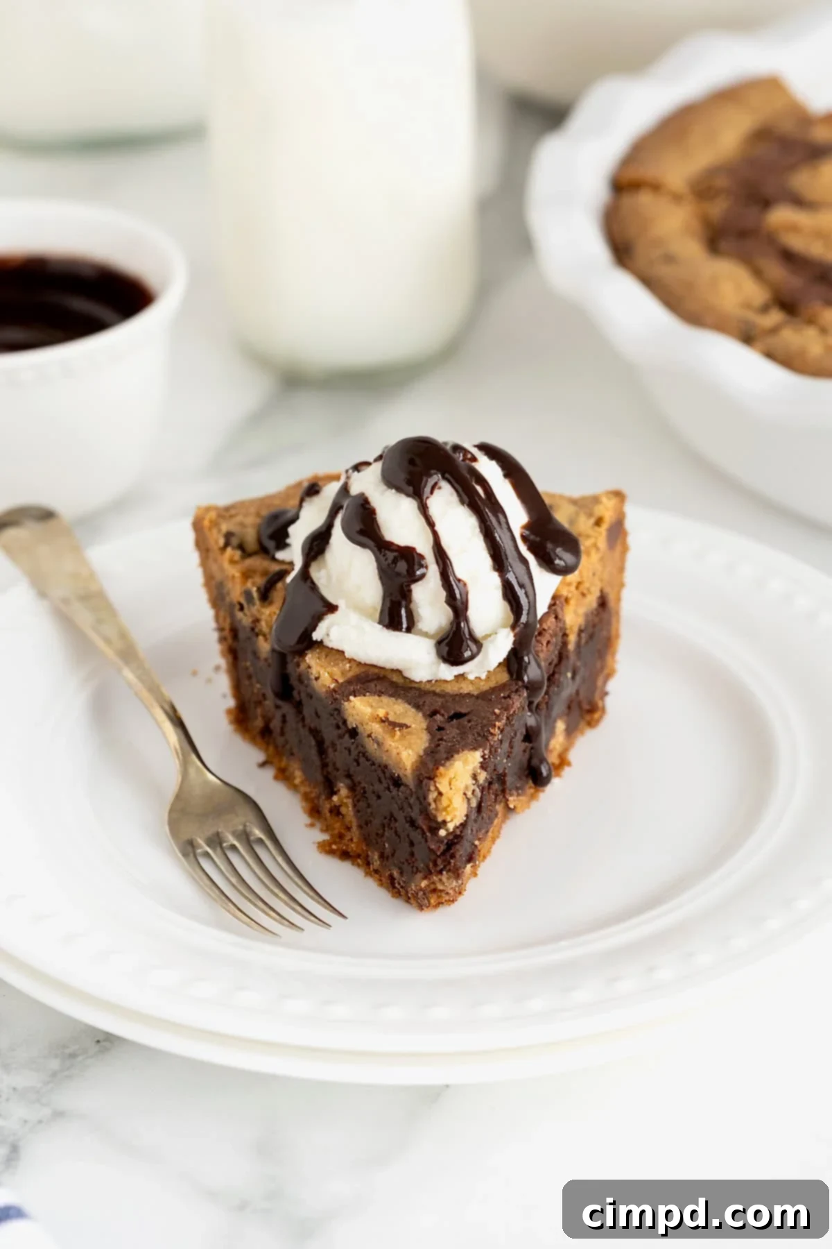 A perfect slice of brookie pie, rich with layers of cookie and brownie, topped with a scoop of vanilla ice cream and chocolate syrup, presented on a small white plate.