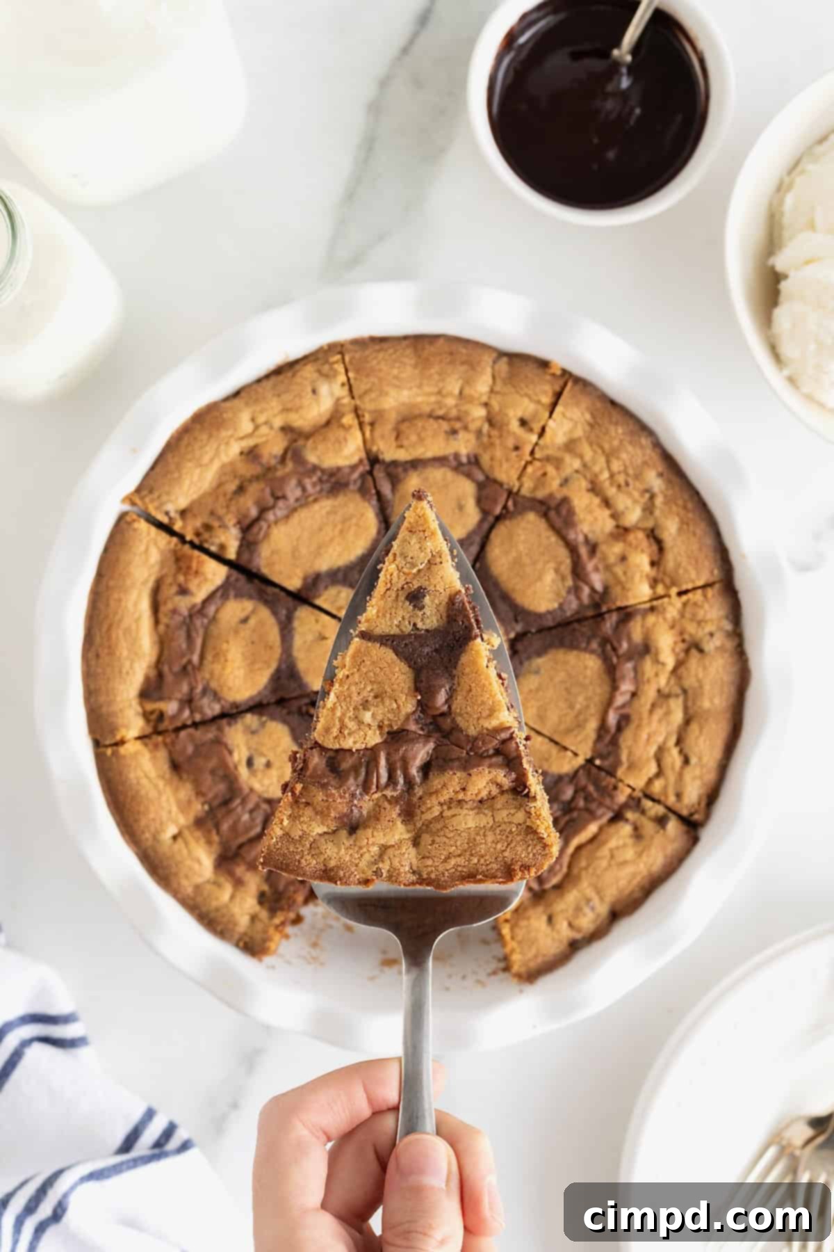 A slice of brookie pie, showing its distinct layers of cookie and brownie, being carefully lifted from a white porcelain pie plate with a metal spatula.