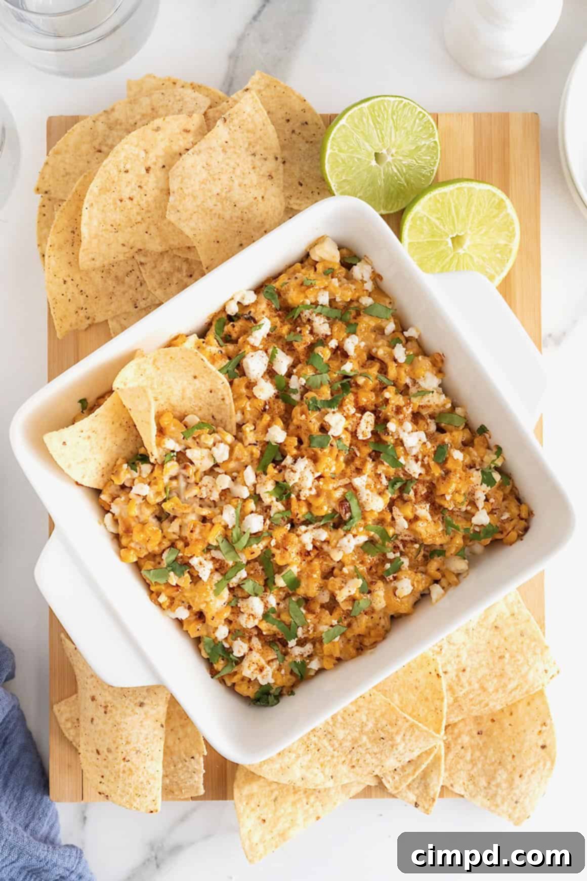 A square white casserole dish of roasted corn elotes dip surrounded by corn chips on a rustic wooden cutting board, ready for serving.