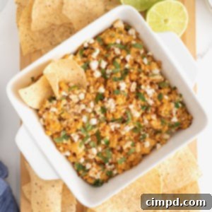 A square white casserole dish of roasted corn elotes dip surrounded by corn chips on a wooden cutting board.