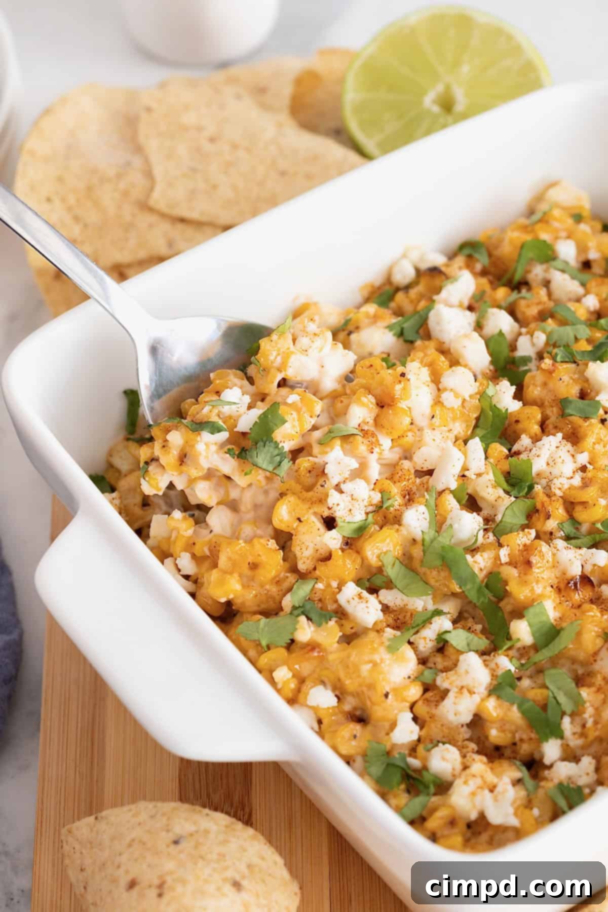 A metal spoon sticks out of a square white casserole dish filled with roasted corn elotes dip, surrounded by corn chips on a wooden cutting board.