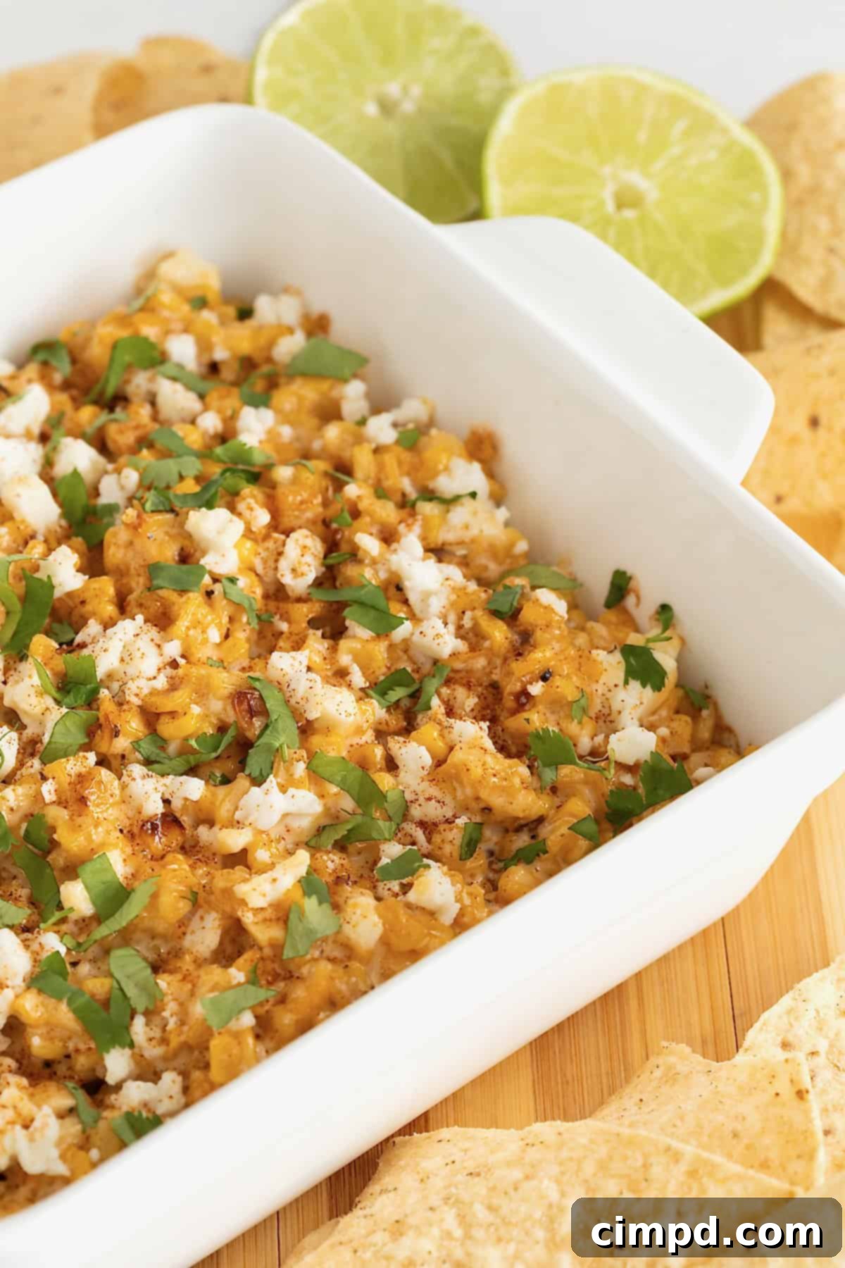 A square white casserole dish of roasted corn elotes dip surrounded by corn chips on a wooden cutting board, ready for serving.