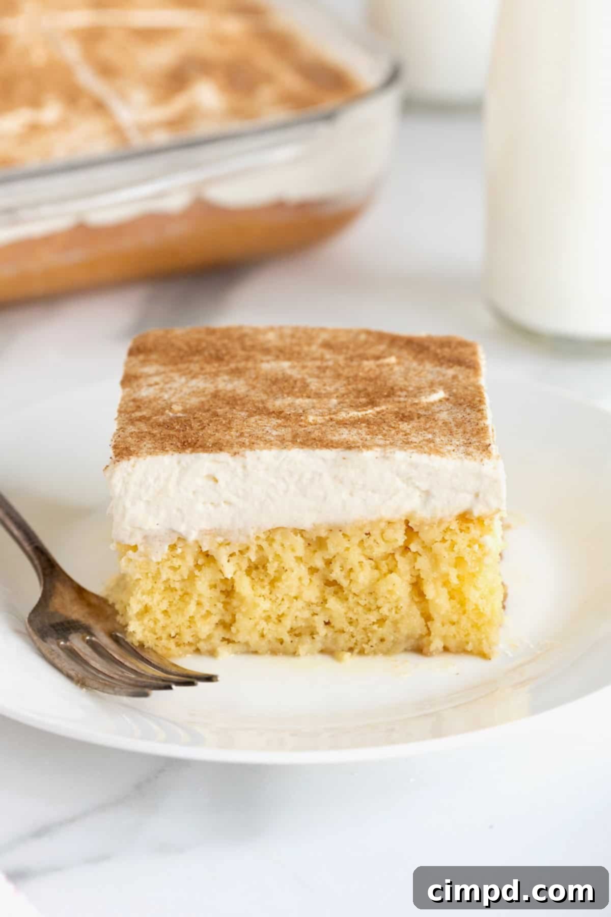 A square of tres leches cake on a white dessert plate with a fork next to it.