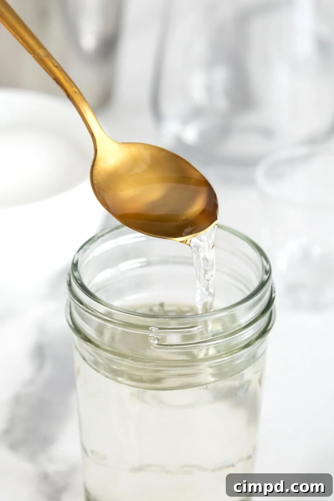 A gold toned spoon pouring simple syrup into a wide mouth glass jar on a white marble counter, highlighting its clear, viscous texture.