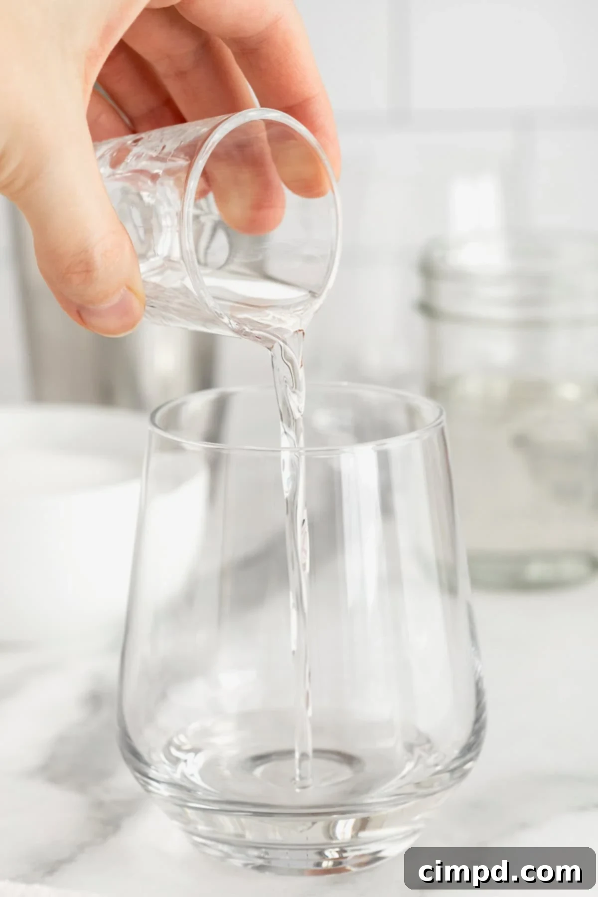 Simple syrup being poured from a small glass pitcher into a cocktail glass on a pristine white marble counter, ready for mixing.
