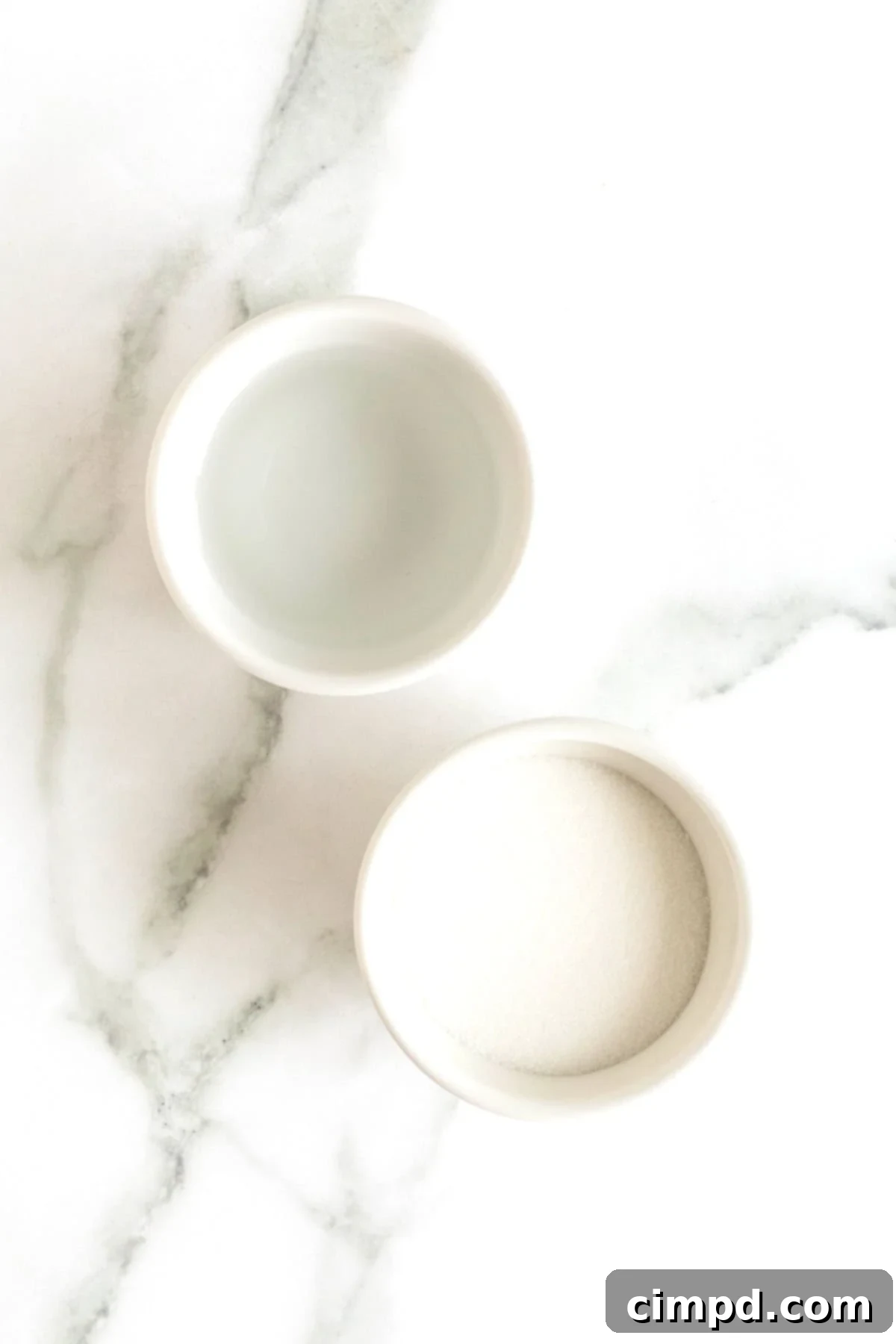 Small white glass dishes containing fresh water and pure granulated sugar, neatly arranged on a white marble counter, ready for making simple syrup.