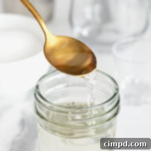 A gold toned spoon pouring simple syrup into a wide mouth glass jar on a white marble counter, showcasing its clear and glossy texture.