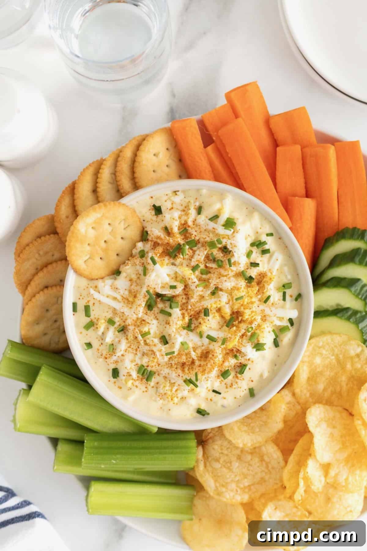 A large white bowl of deviled egg dip surrounded by crackers, carrot sticks, cucumber slices, potato chips and celery sticks on a large round platter.