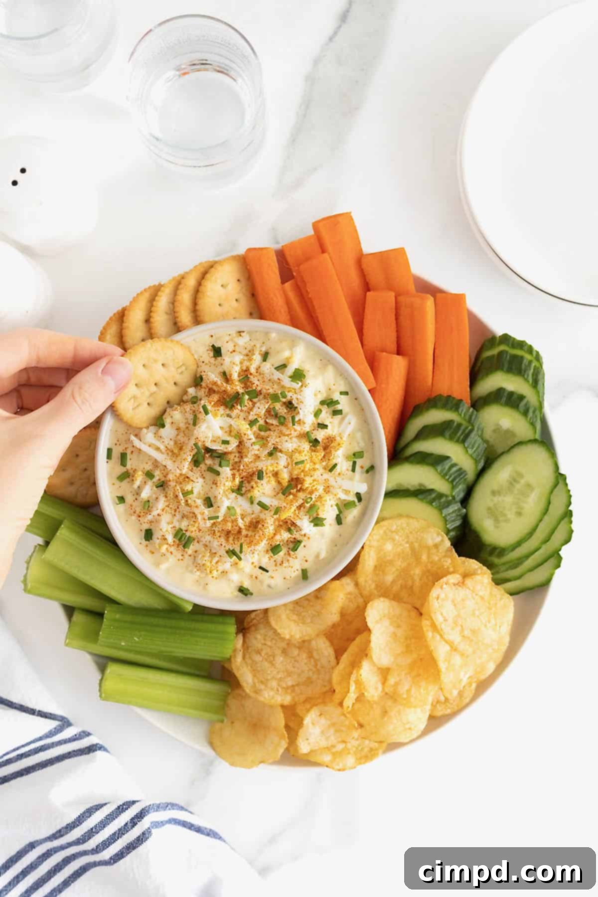 A hand dipping a round butter cracker into a bowl of Deviled Egg Dip surrounded by fresh vegetables, crackers and potato chips.