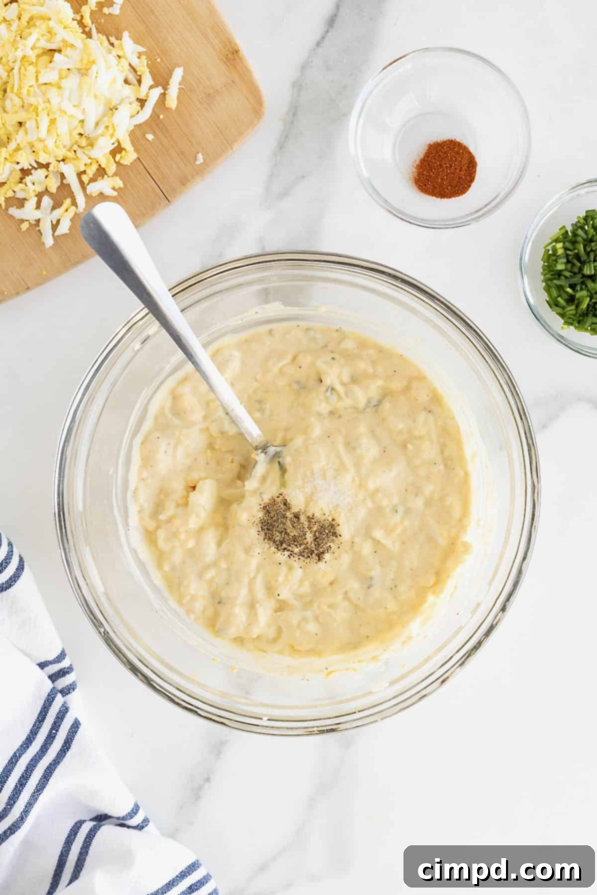 Salt and pepper being added to a large glass mixing bowl of deviled egg dip.