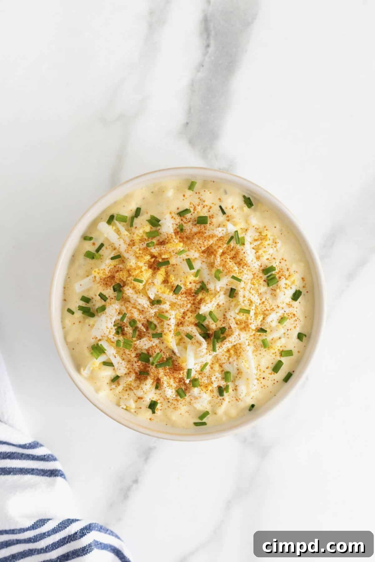 A white serving bowl of deviled egg dip garnished with paprika, chives and grated egg pieces on a white marble counter.