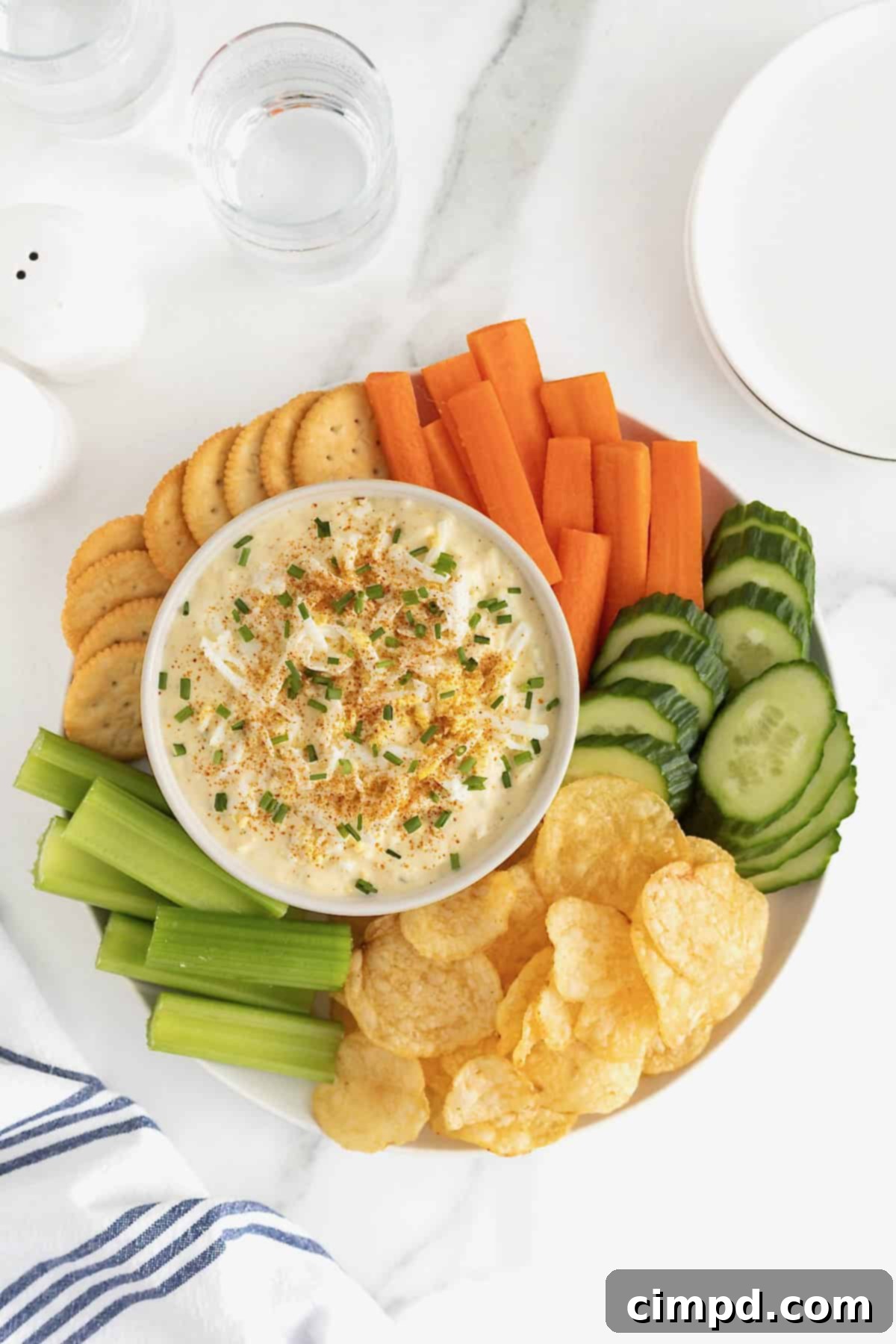 A large white bowl of deviled egg dip surrounded by crackers, carrot sticks, cucumber slices, potato chips and celery sticks on a large round platter.