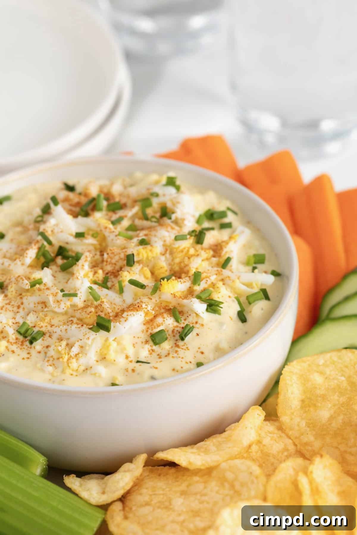 A large white bowl of deviled egg dip surrounded by crackers, carrot sticks, cucumber slices, potato chips and celery sticks on a large round platter.