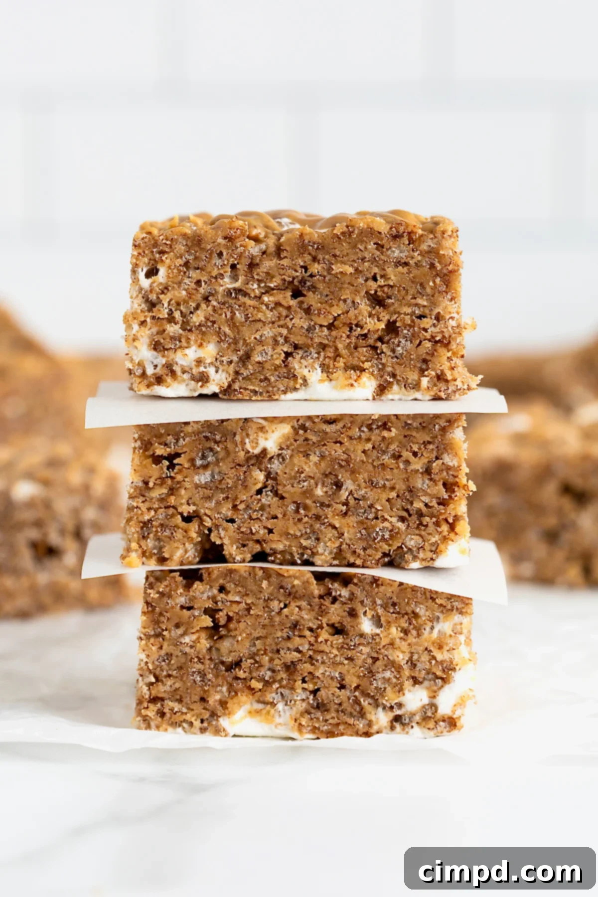 A stack of three perfectly cut Chocolate Peanut Butter Rice Krispies Treats, each separated by parchment paper, sits on a clean white marble counter, highlighting their appealing layers.