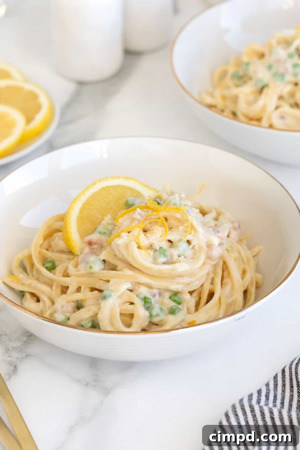 A gold rimmed white bowl of linguini with a creamy white sauce, vibrant green peas, and crispy pancetta, presented on a elegant white marble countertop.