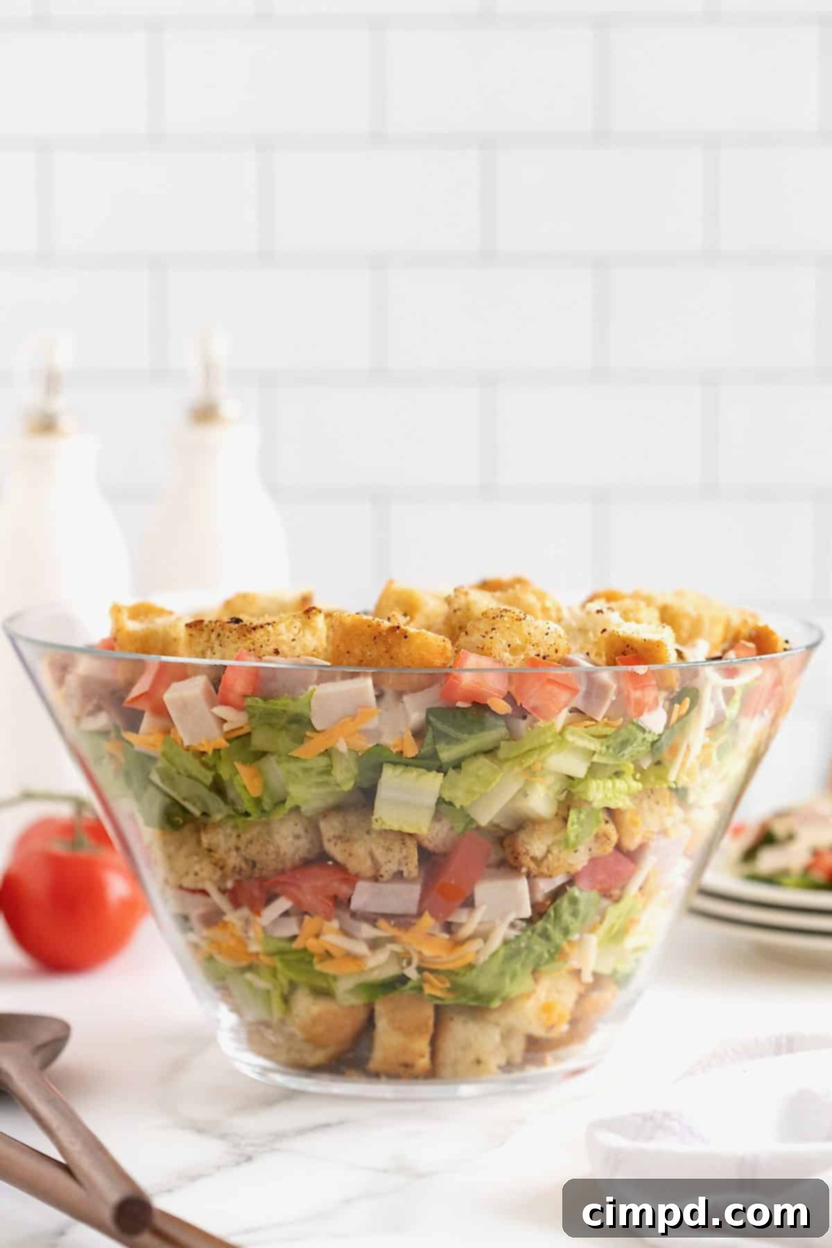 Club Salad Parfait 11 A large clear glass salad bowl filled with layers of croutons, salad mix, shredded cheese, bacon, ham and tomatoes.
