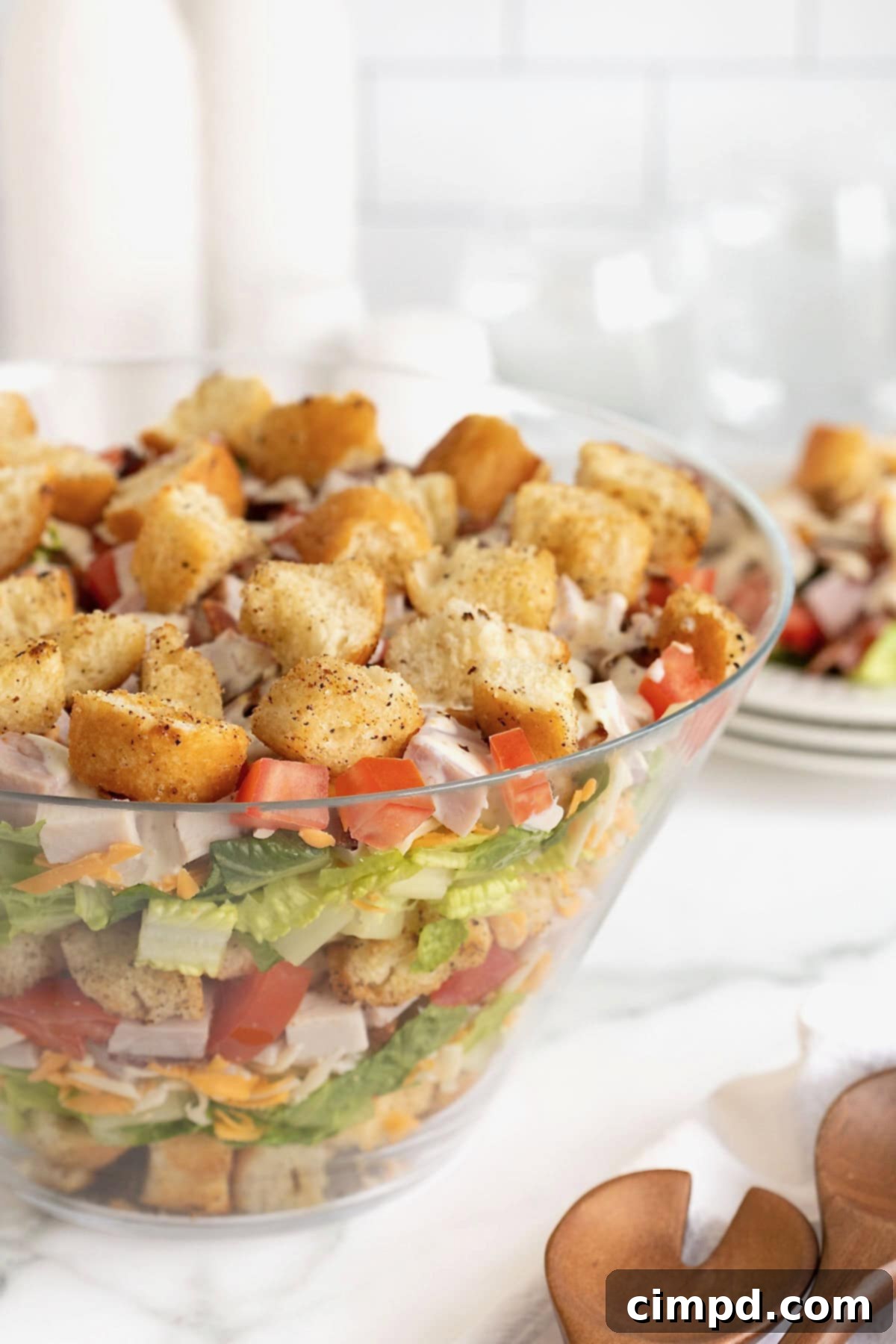 Club Salad Parfait 4 A large clear glass salad bowl filled with layers of croutons, salad mix, shredded cheese, bacon, ham and tomatoes.