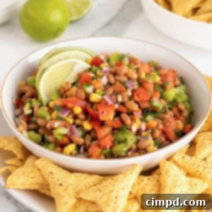 A top-down view of colorful Baked Bean Cowboy Caviar in a white bowl, surrounded by corn tortilla chips on a white platter, ready for a party.