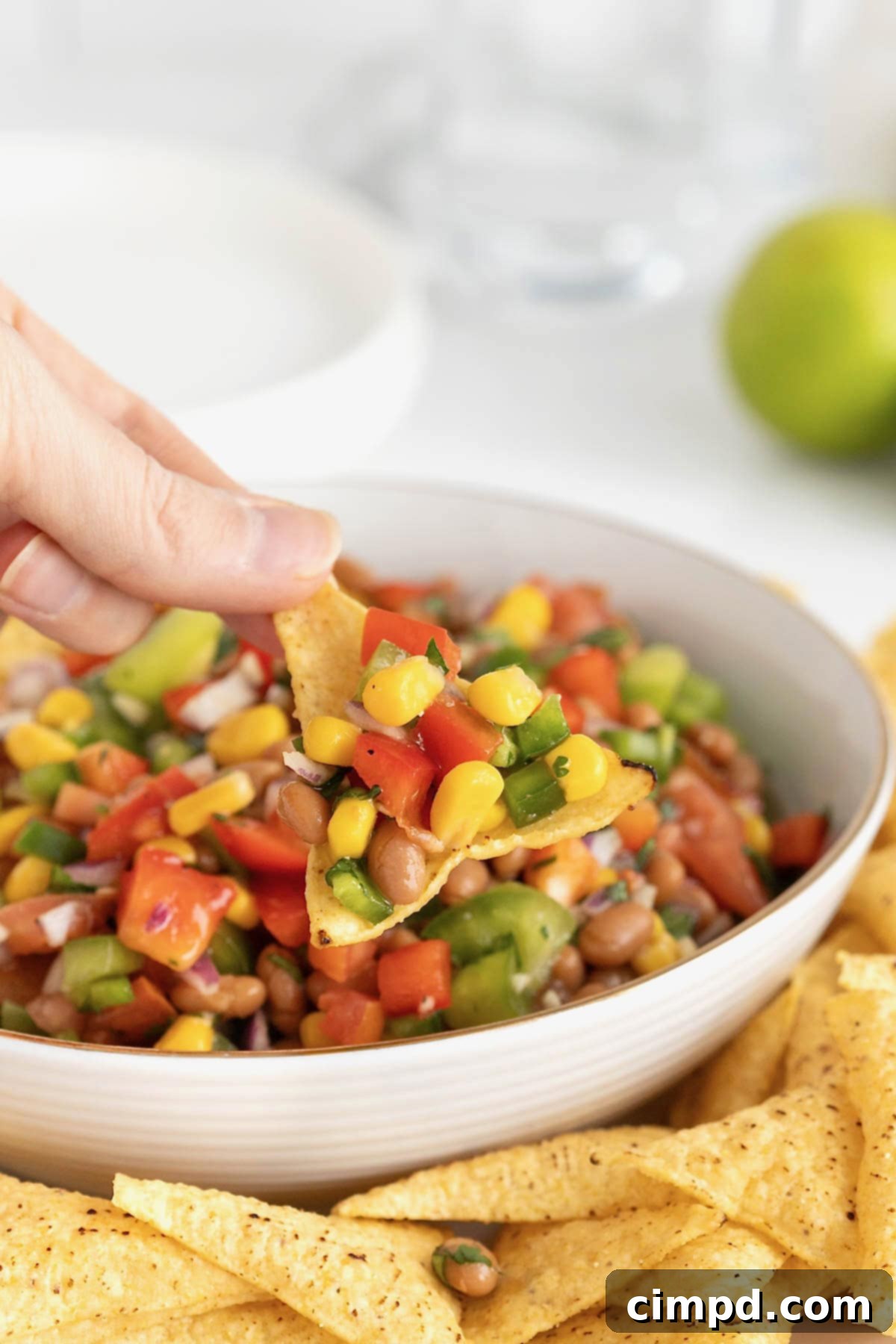 A corn tortilla chip laden with a generous scoop of Baked Bean Cowboy Caviar, held above a rustic white bowl brimming with the colorful dip.