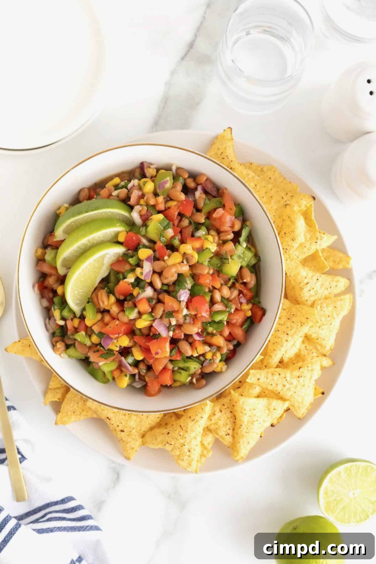 A pristine white bowl overflowing with colorful Cowboy Caviar, surrounded by a scattering of golden corn tortilla chips on a matching white platter, indicating it's ready for a party.