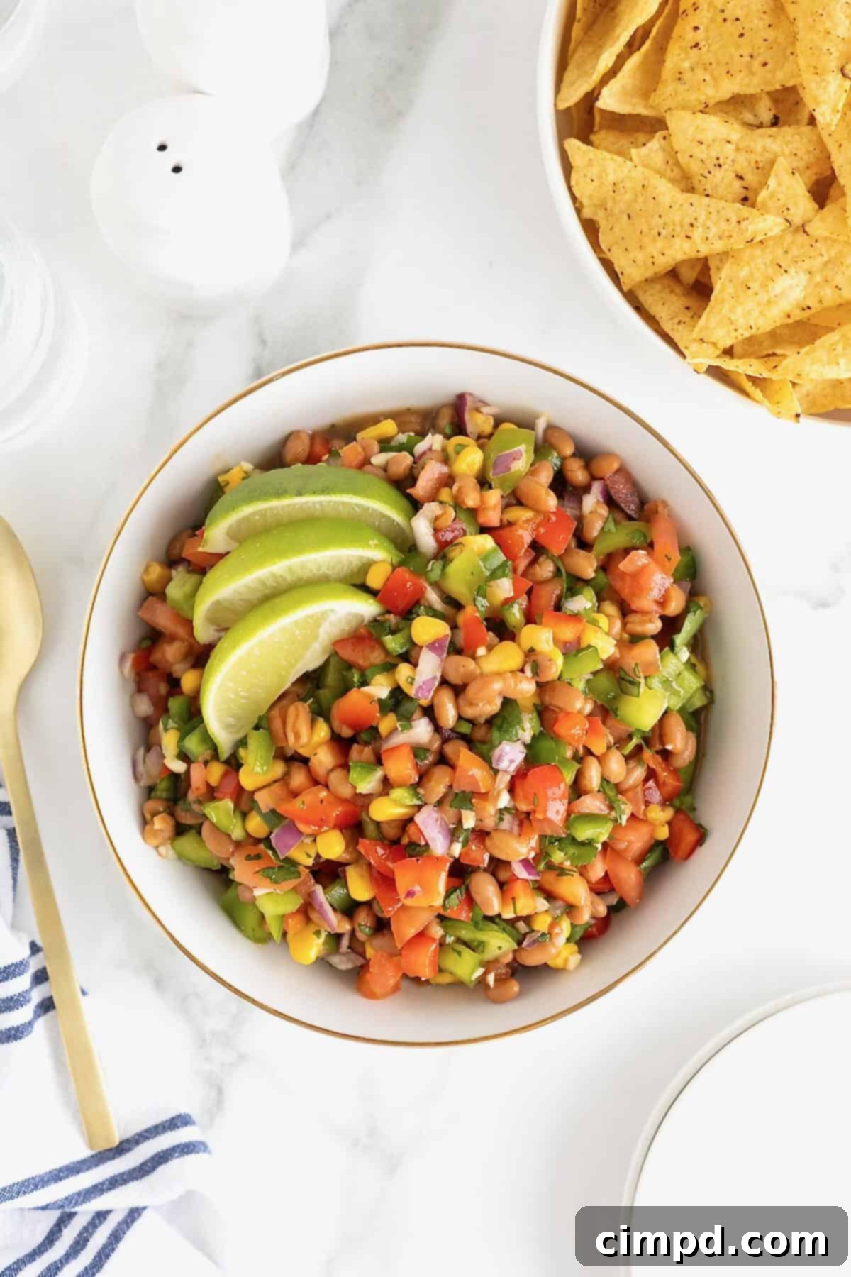A large gold-rimmed white serving bowl of Baked Bean Cowboy Caviar, garnished with two slices of lime, ready to be enjoyed as a refreshing dip.