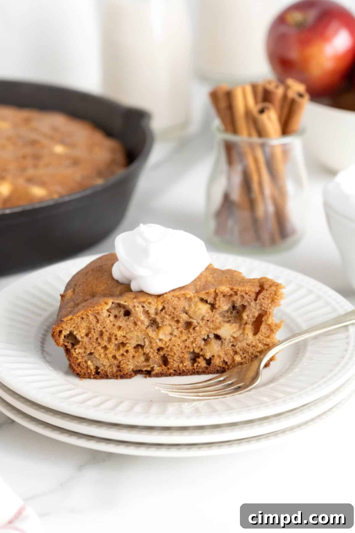 A golden-brown slice of apple skillet cake, generously topped with a dollop of fluffy whipped cream, resting on a stack of three elegant white fluted dessert plates. A polished metal fork is positioned neatly beside the cake, ready to indulge.