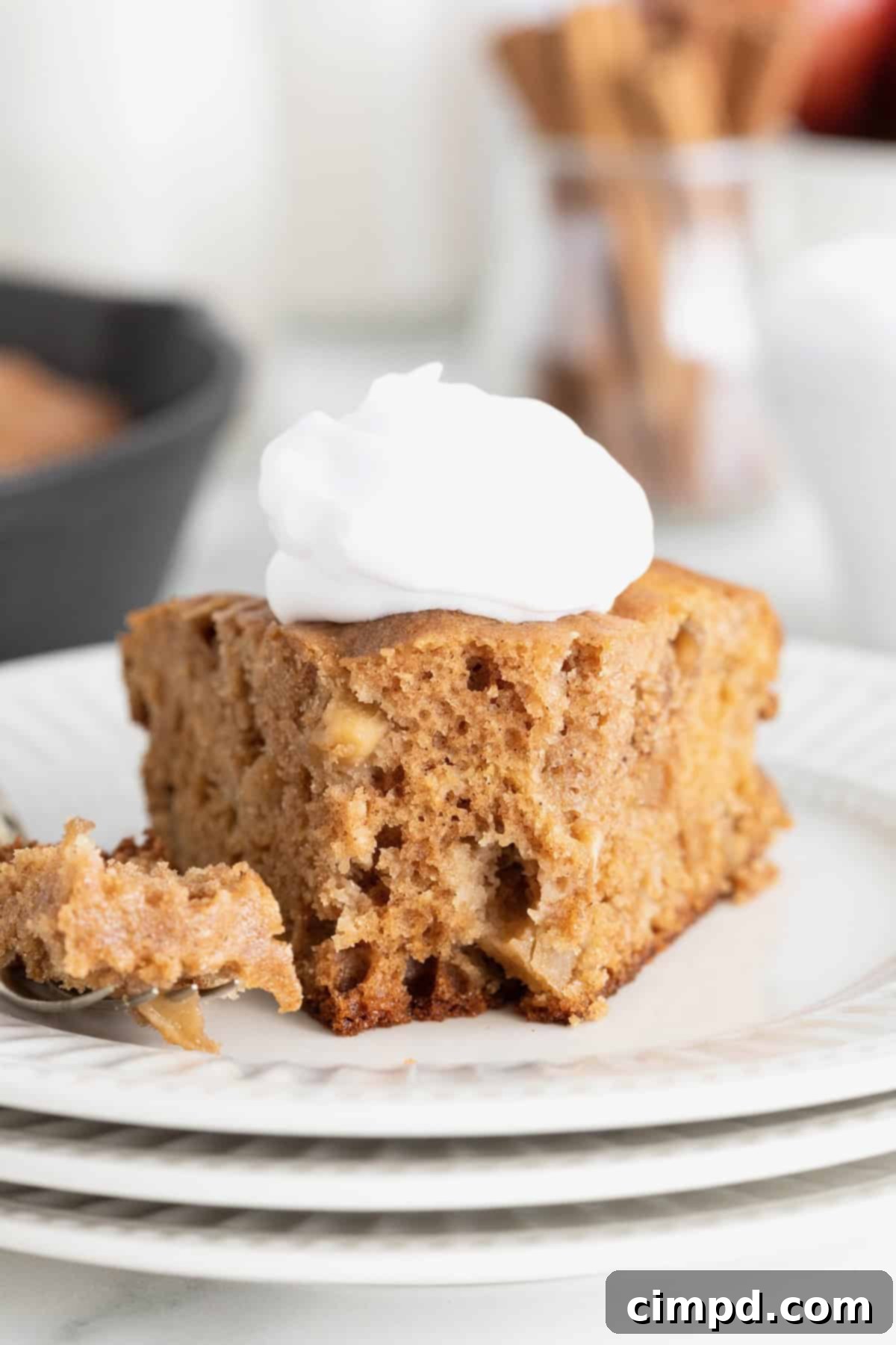 A beautifully presented slice of apple skillet cake, adorned with a dollop of fresh whipped cream, sits gracefully on a stack of three white fluted dessert plates. A silver fork rests nearby, hinting at the delightful experience awaiting.