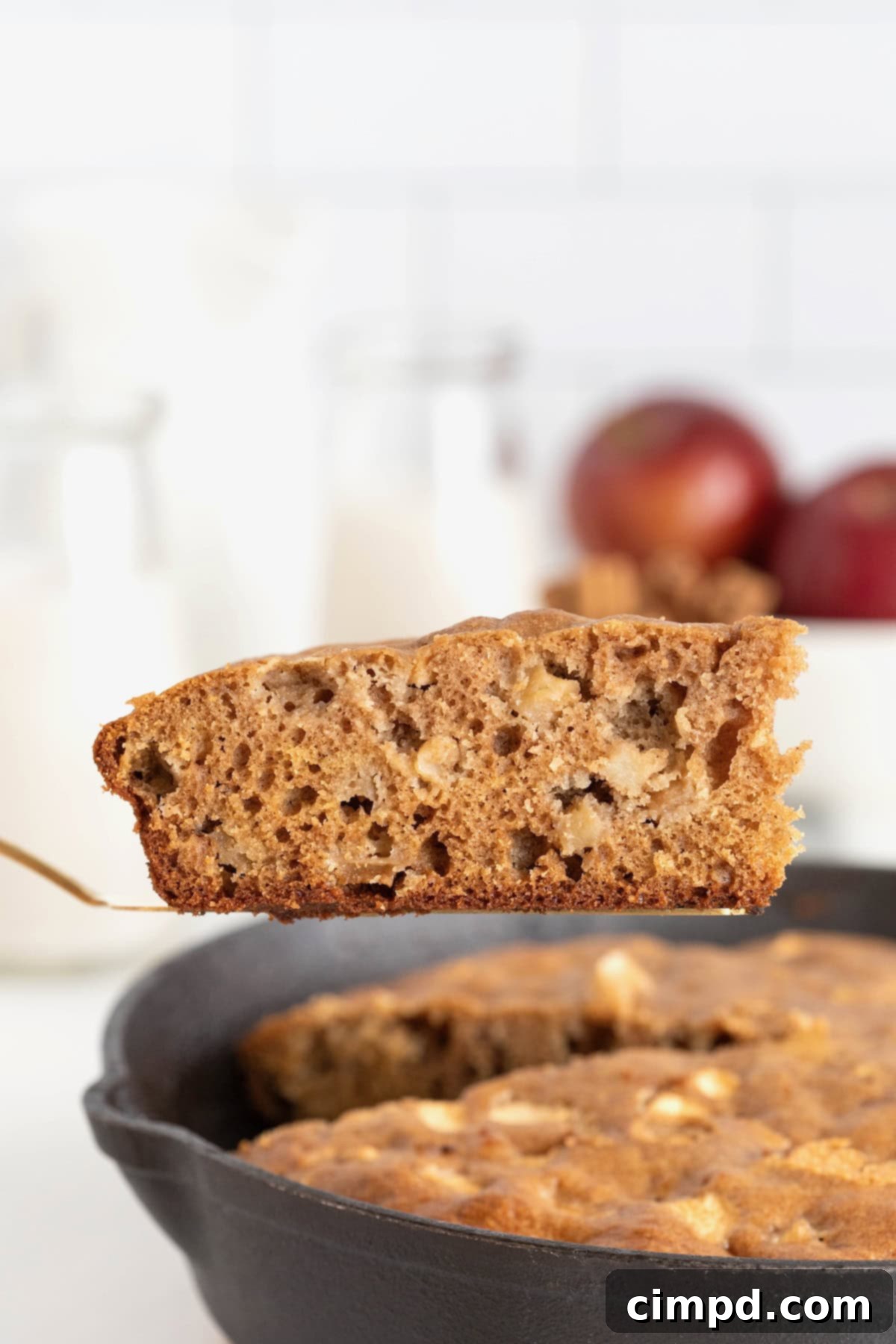 A freshly baked slice of apple cake is being lifted out of a warm cast iron skillet with a spatula, showcasing its tender texture and golden crust.