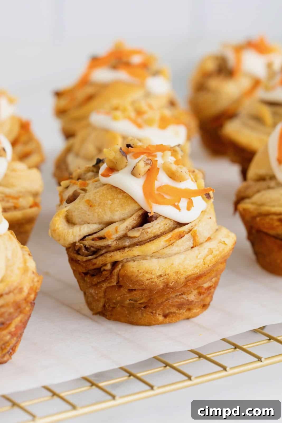 Flaky Spiced Carrot Cruffins 2 Nine perfectly baked Carrot Cake Cruffins, each generously topped with creamy white cream cheese frosting, sprinkled with chopped walnuts, and adorned with delicate ribbons of fresh shredded carrot, arranged on a parchment-lined cooling rack. The golden-brown pastry layers are visible, promising a flaky texture.