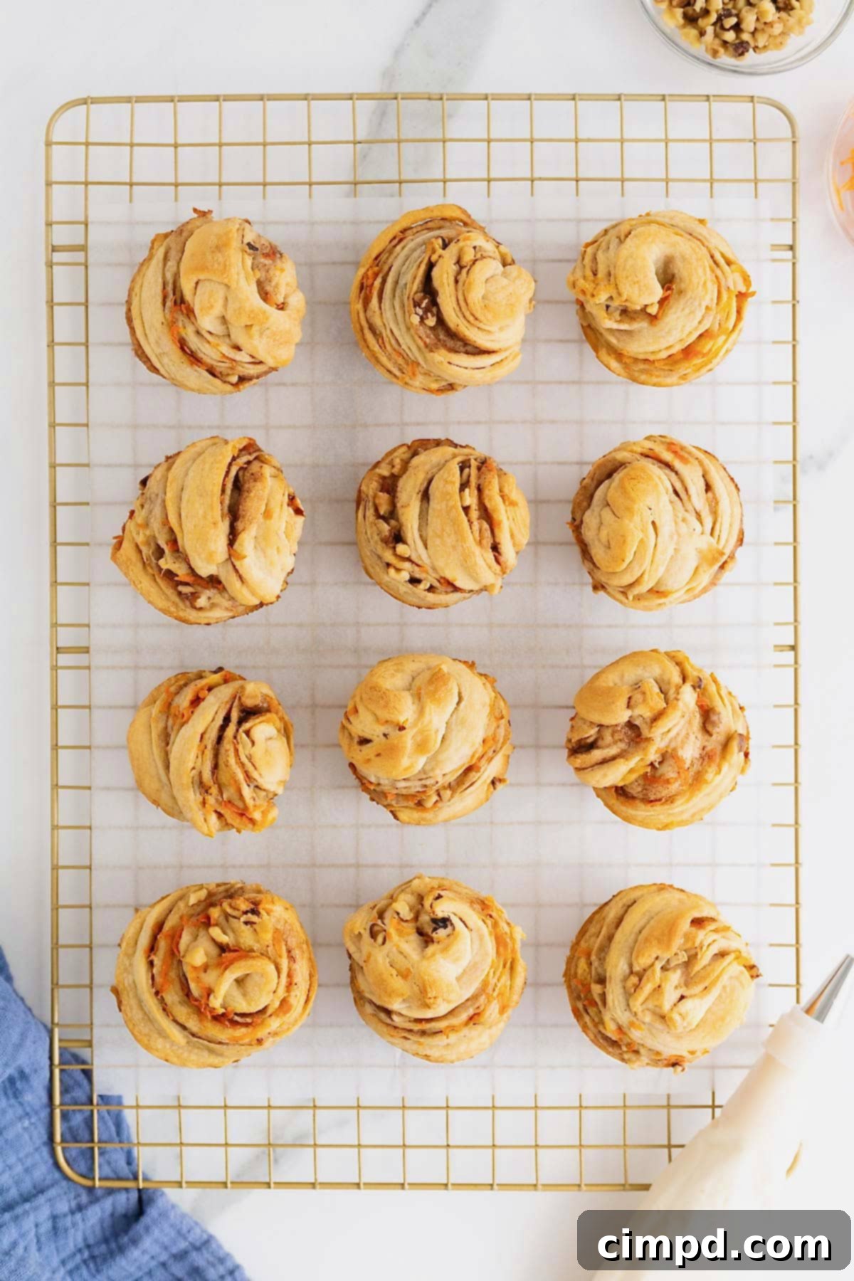 Flaky Spiced Carrot Cruffins 11 Twelve beautifully frosted Carrot Cake Cruffins, each topped with a swirl of cream cheese icing, are neatly arranged on a parchment-lined cooling rack, ready to be served.