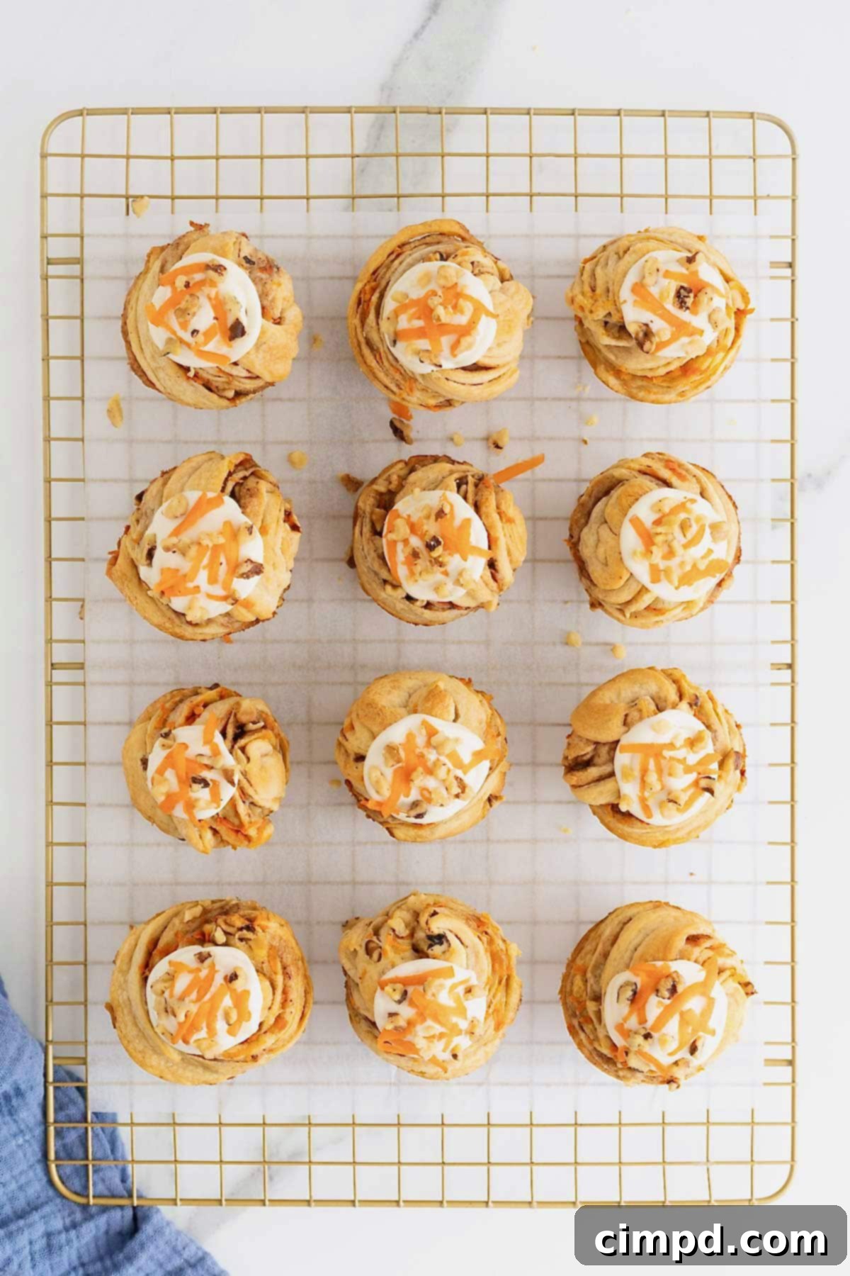 Flaky Spiced Carrot Cruffins 12 A dozen freshly baked Carrot Cake Cruffins, elegantly arranged on a gold-toned cooling rack lined with parchment paper, each crowned with creamy frosting, walnuts, and shredded carrot. They are ready to be enjoyed.