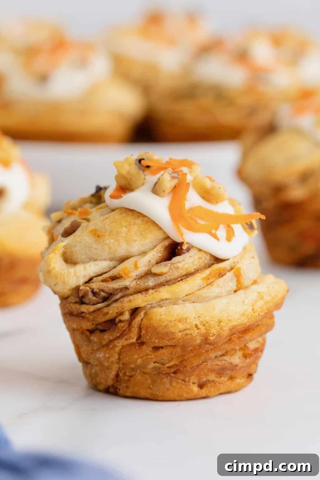 Flaky Spiced Carrot Cruffins 4 A single, beautifully crafted Carrot Cake Cruffin, topped with a dollop of cream cheese frosting and a sprinkle of walnuts, rests on a pristine white marble counter. Its flaky layers are visible, highlighting the artisanal quality.