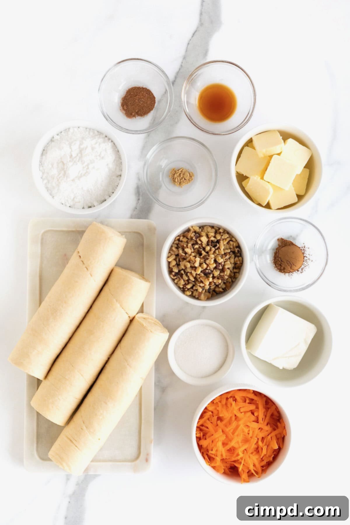 Flaky Spiced Carrot Cruffins 6 A collection of ingredients for Carrot Cake Cruffins, including shredded carrots, spices, butter, cream cheese, and walnuts, neatly arranged in small glass dishes on a white marble counter, ready for preparation.