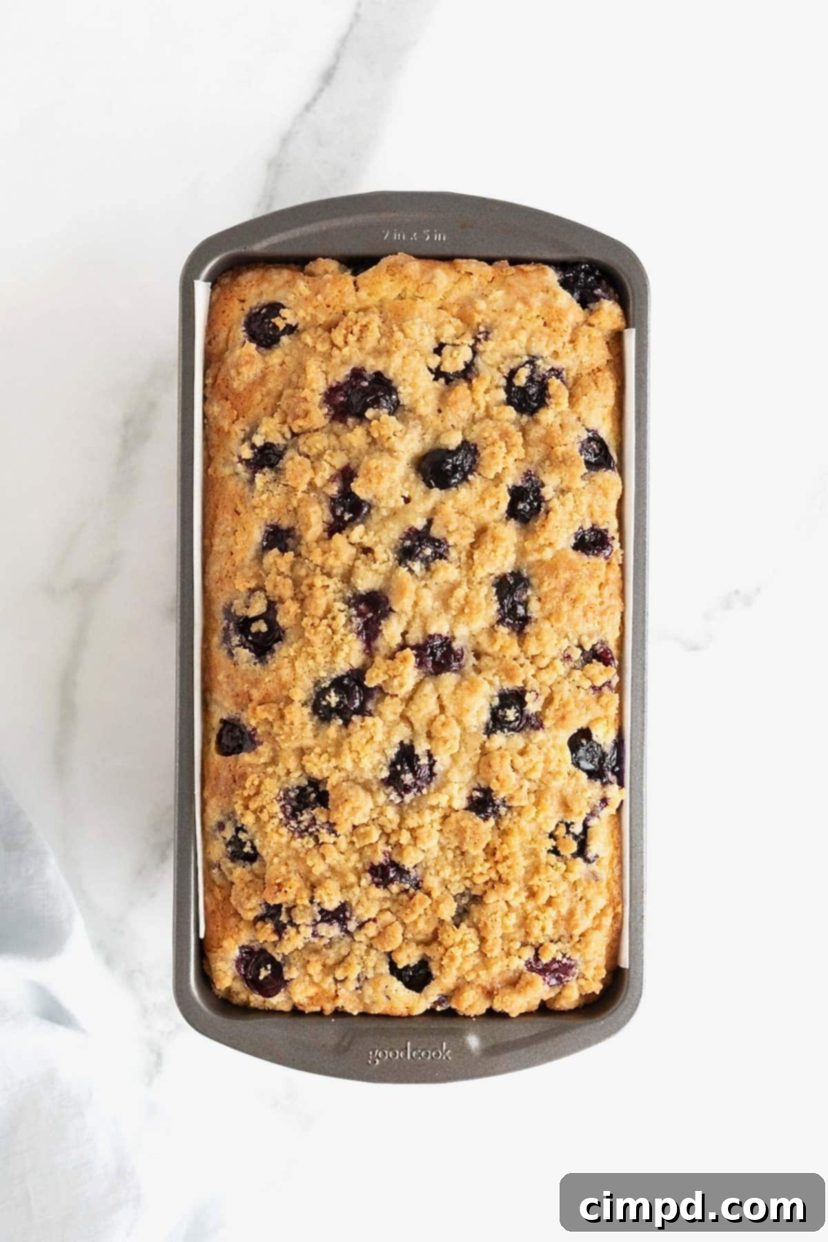 A loaf of lemon blueberry bread in a dark aluminum loaf pan on a white marble counter.