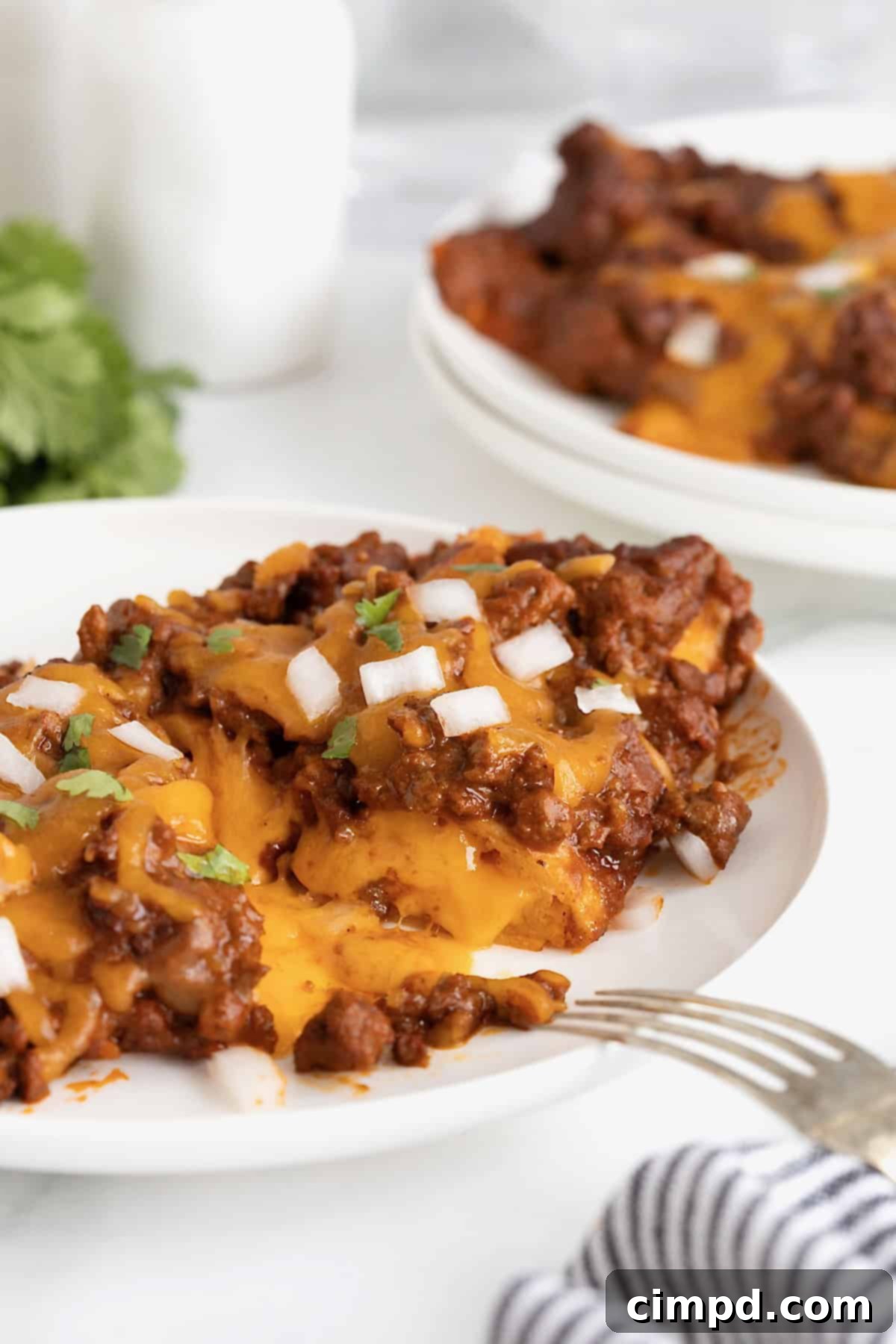 A fork piercing into a delicious Chili Con Carne Enchilada on a white plate, showcasing the rich sauce and melted cheese.