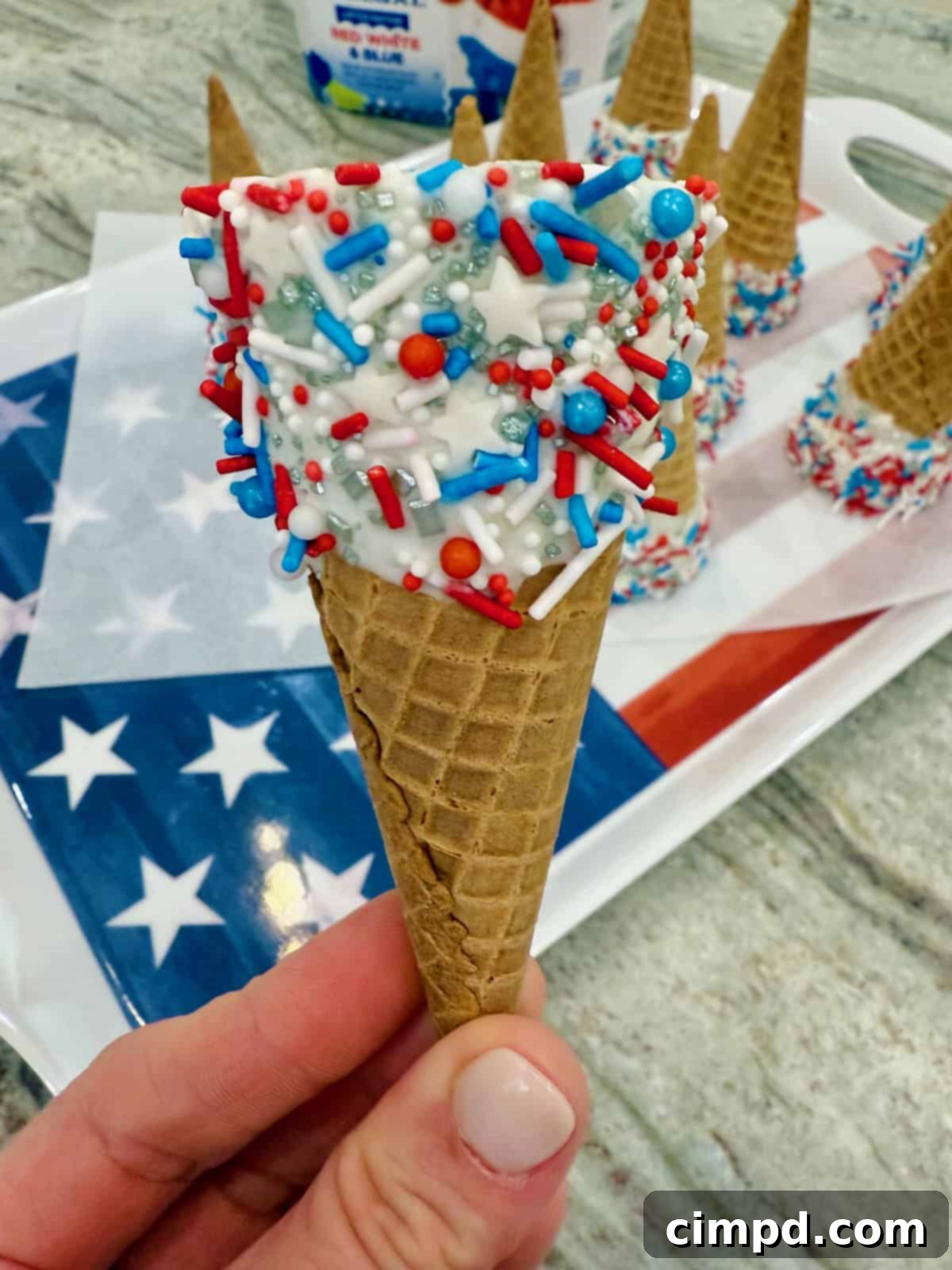 Simple Star-Spangled Bites 4 A crisp sugar cone artfully decorated with a white vanilla candy coating and a vibrant mix of red, white, and blue sprinkles, ready for ice cream.