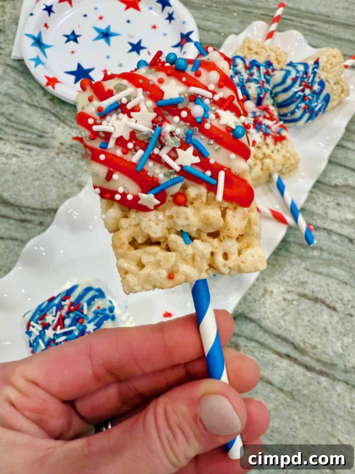 Simple Star-Spangled Bites 6 A festive Rice Krispies treat on a blue striped straw, half-dipped in white candy coating, drizzled with red icing, and covered in colorful patriotic sprinkles.