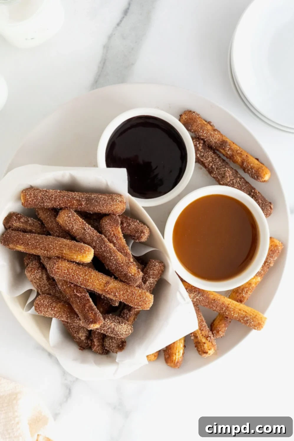 Guilt-Free Baked Churros 11 A parchment lined bowl of baked churros next to a small white dish of chocolate sauce and a small white dish of caramel sauce on a large white platter.